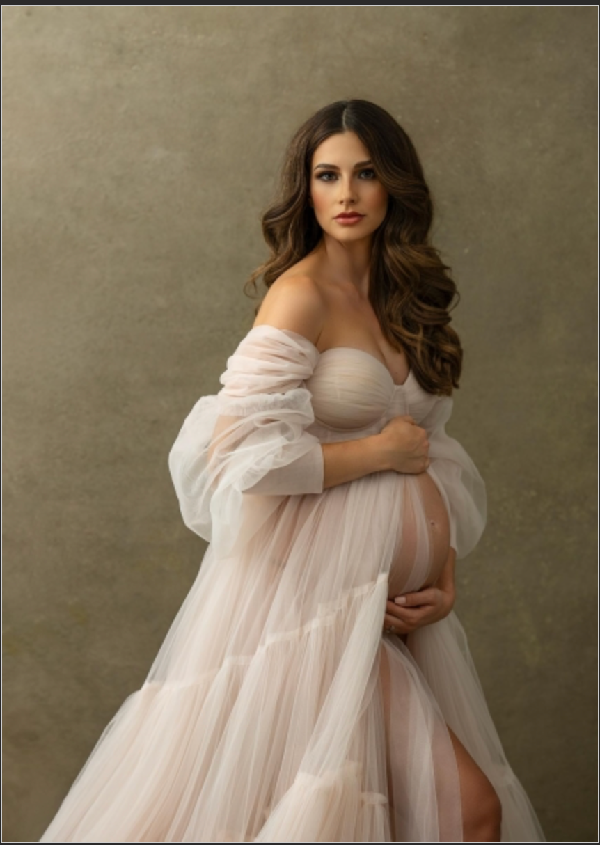 Elegant pregnant woman in a sheer off-shoulder blush gown posing for a glamorous maternity portrait.