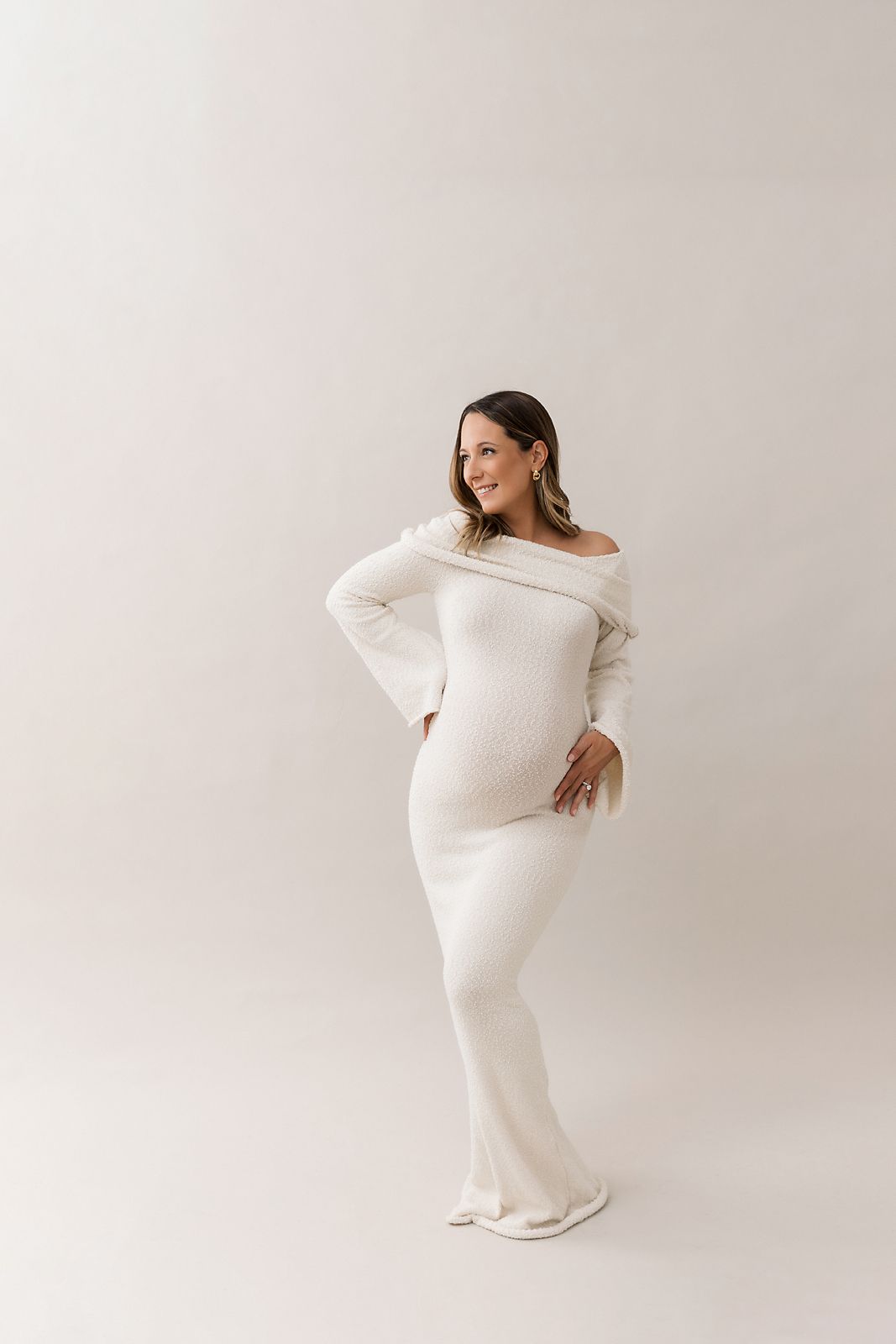 Pregnant woman in white off-shoulder ribbed maxi dress posing gracefully in a bright studio setting.