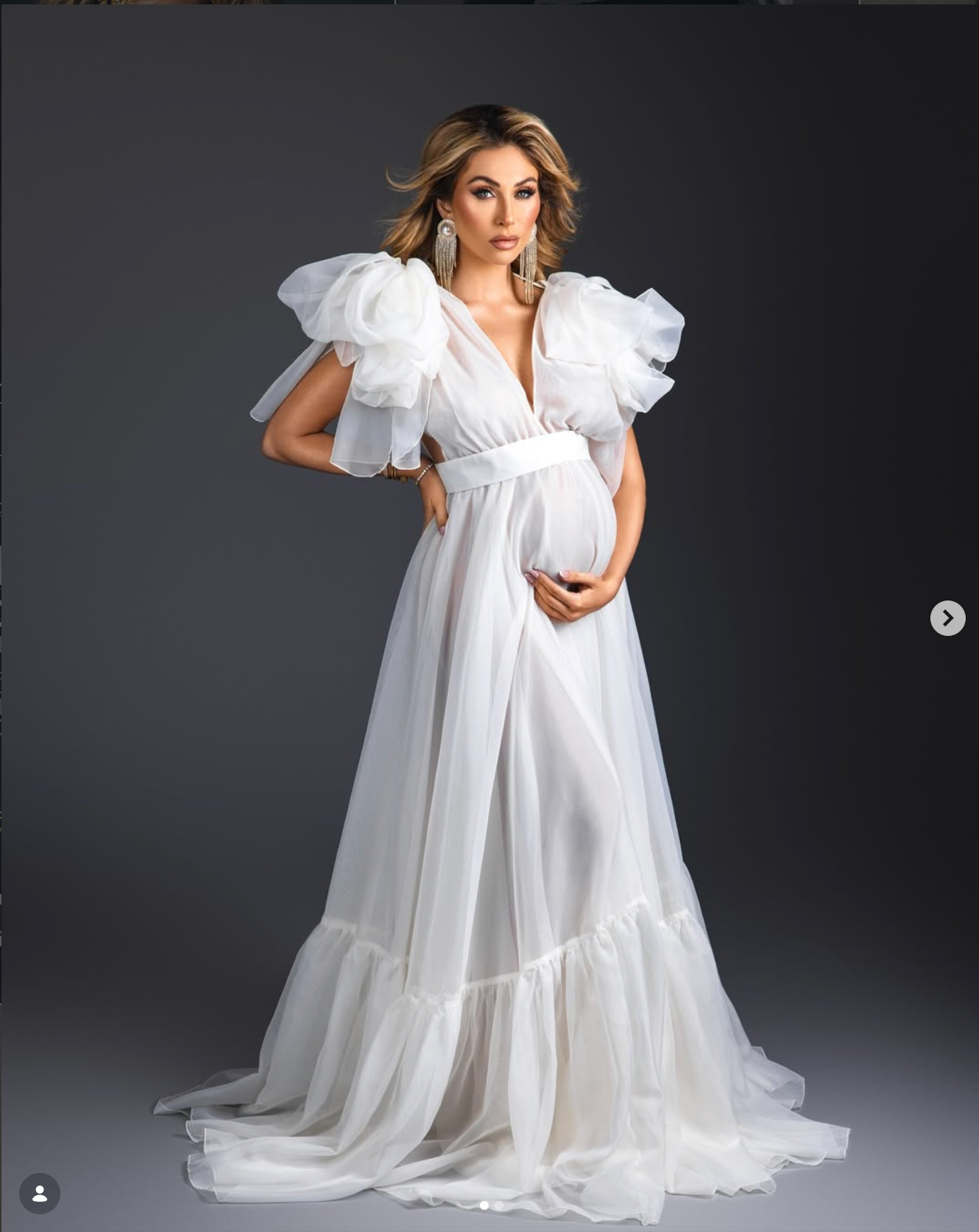 Pregnant woman in elegant white ruffled tulle maternity gown posing gracefully against a dark studio background.