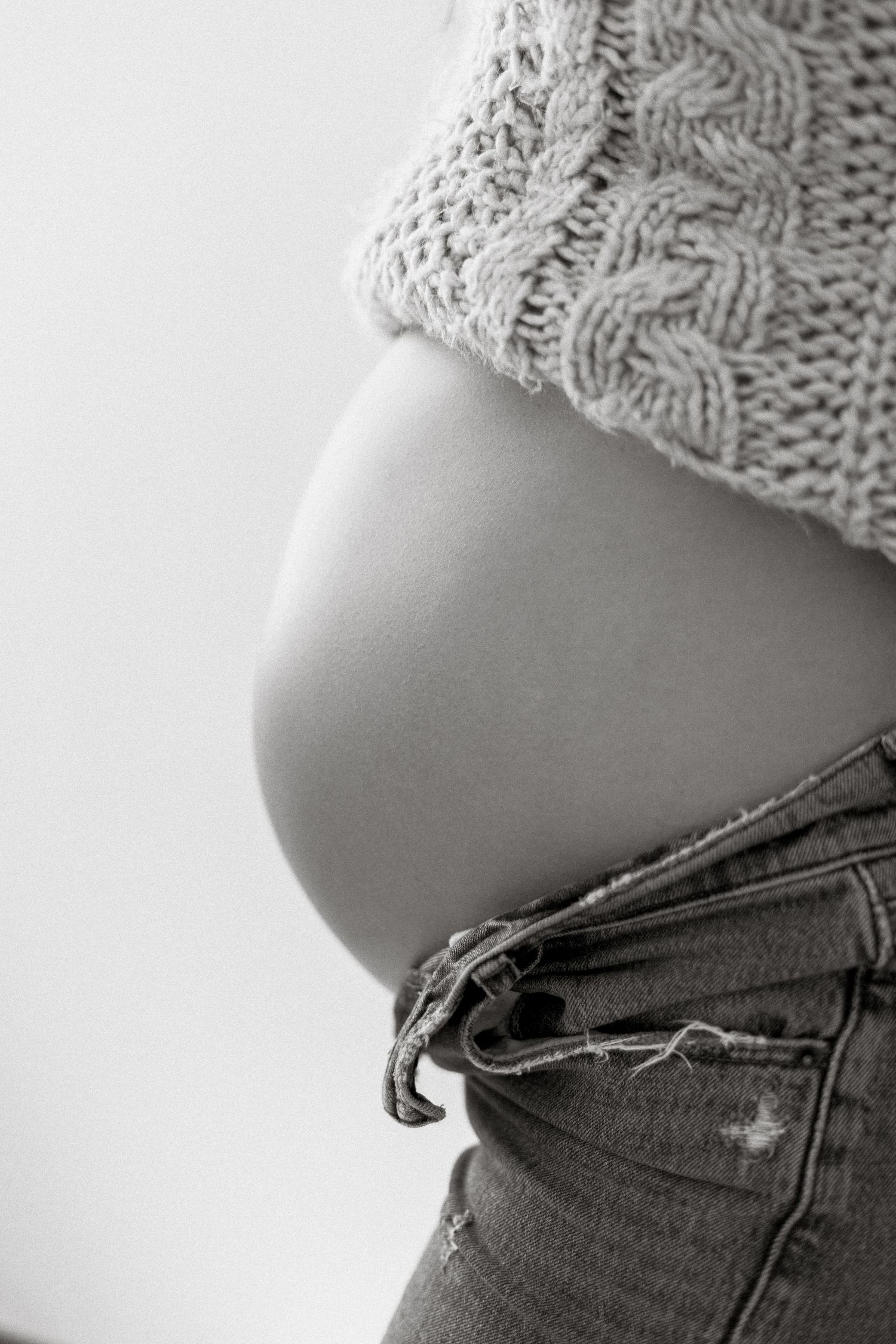 Black and white photo of a pregnant belly showing beneath a knit sweater with distressed denim jeans.