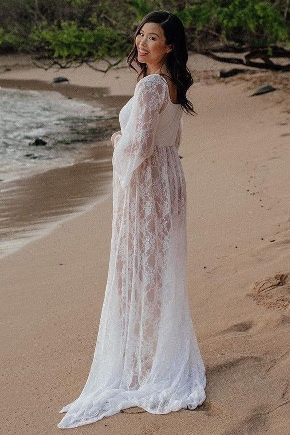 Woman in white lace maternity gown standing on sandy beach, looking back over shoulder with lush greenery behind.