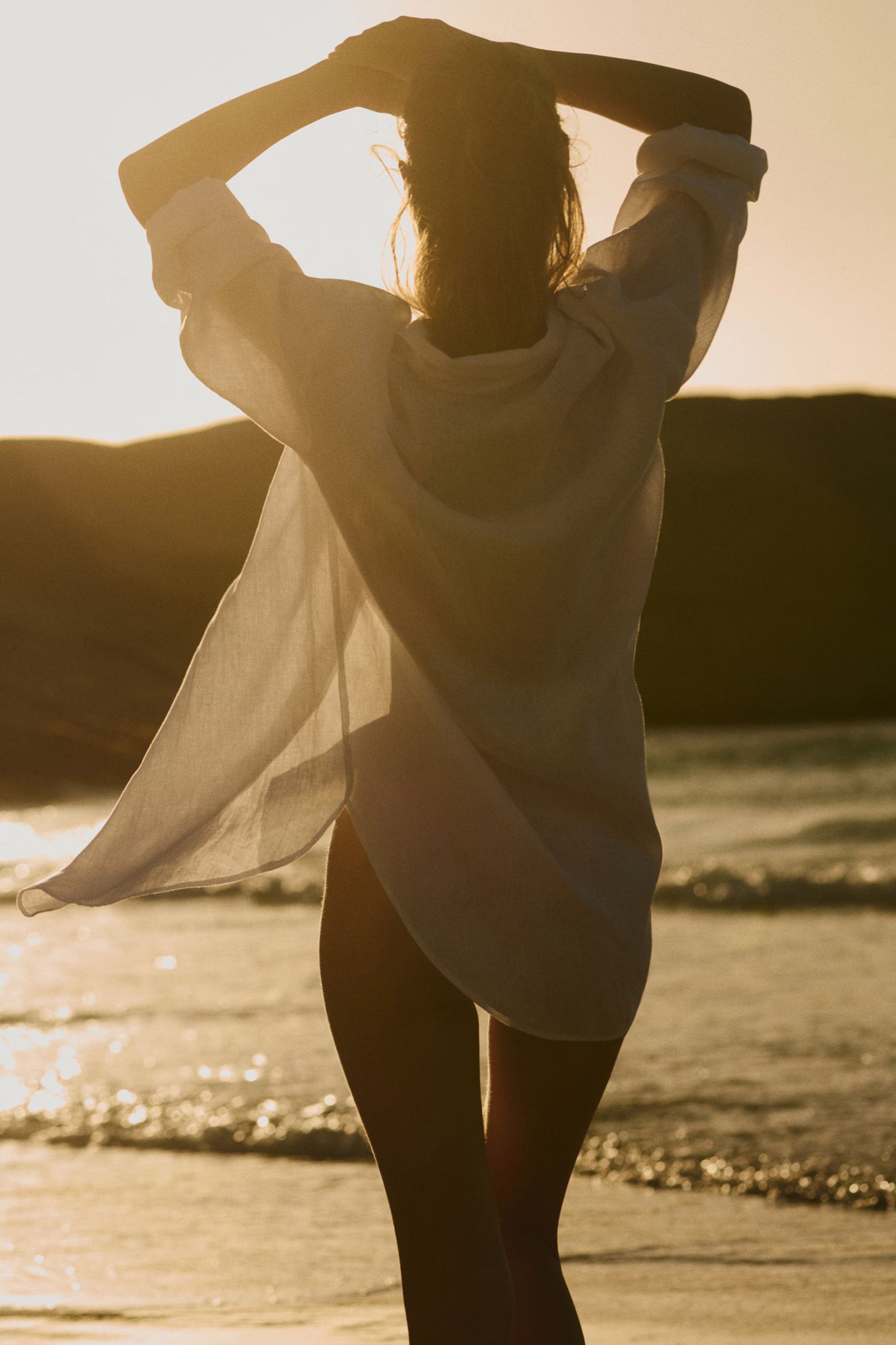 Silhouette of woman in sheer white shirt at golden sunset beach, arms raised.