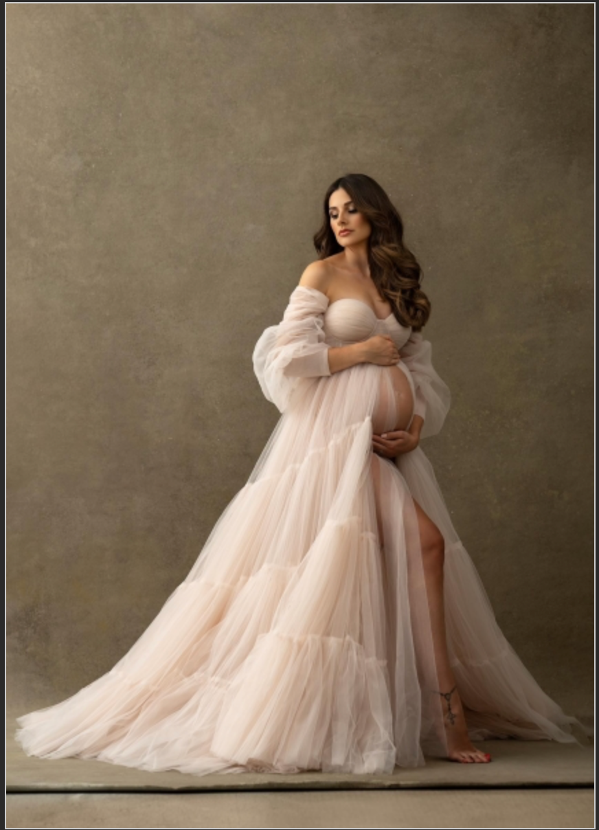 Pregnant woman in elegant off-shoulder blush tulle maternity gown posing gracefully in a professional studio setting.