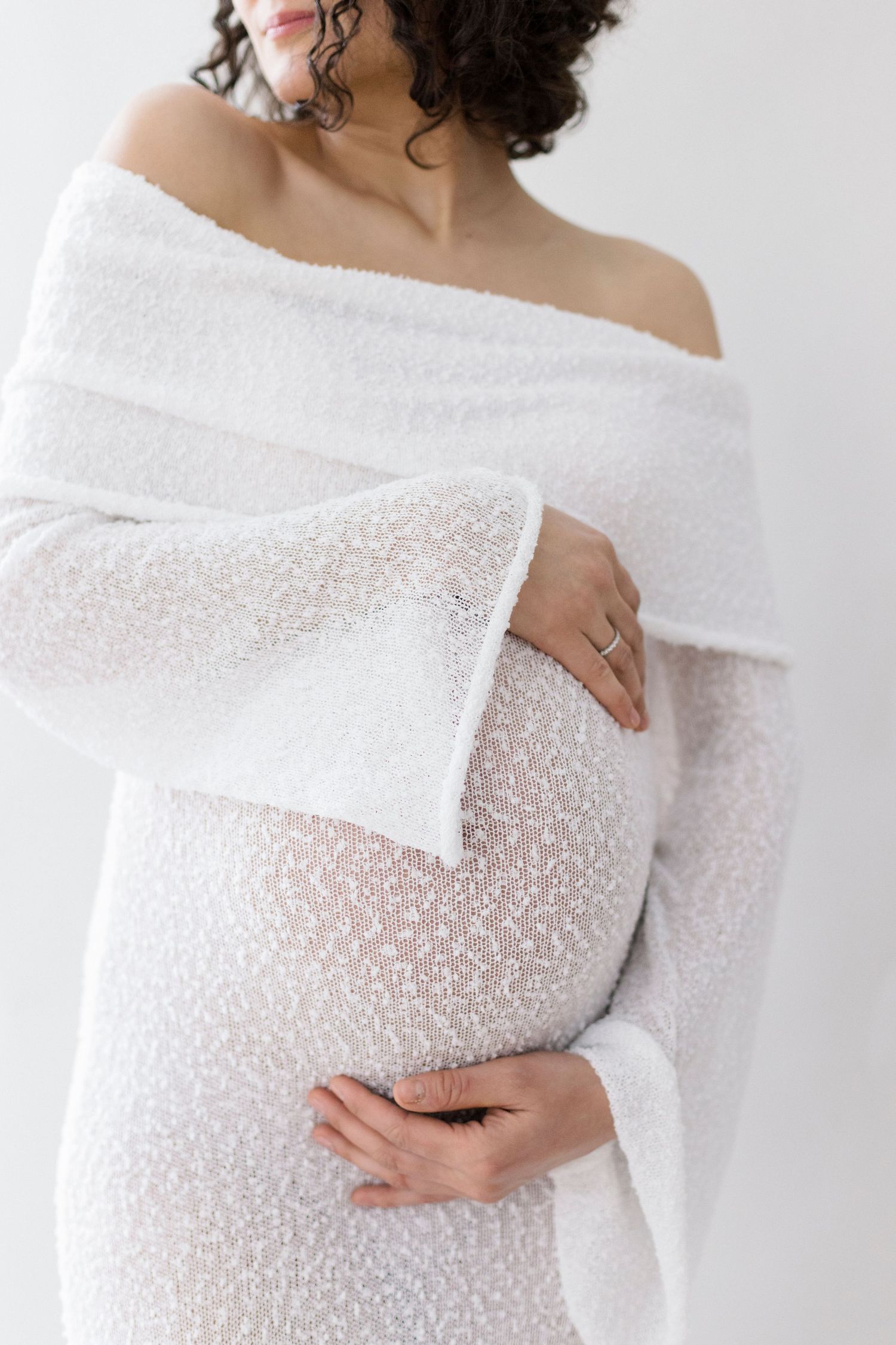 Pregnant woman in white off-shoulder knit dress cradling baby bump, maternity photography on white background.