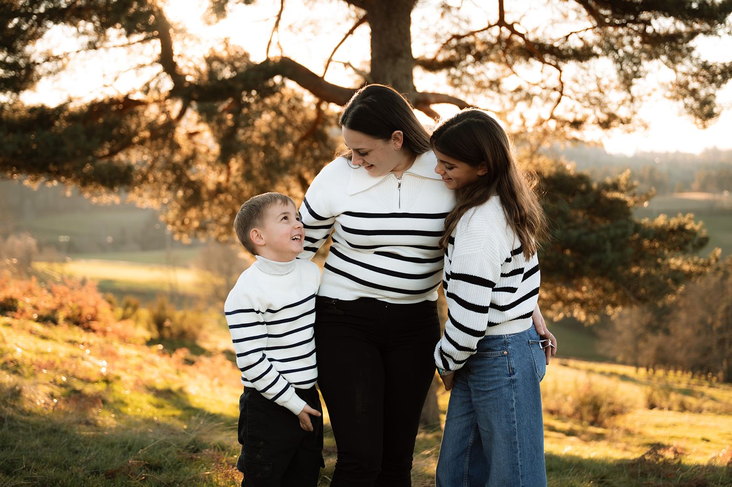 A couple in striped shirts and jeans embrace during a golden sunset photoshoot in a rustic outdoor setting.