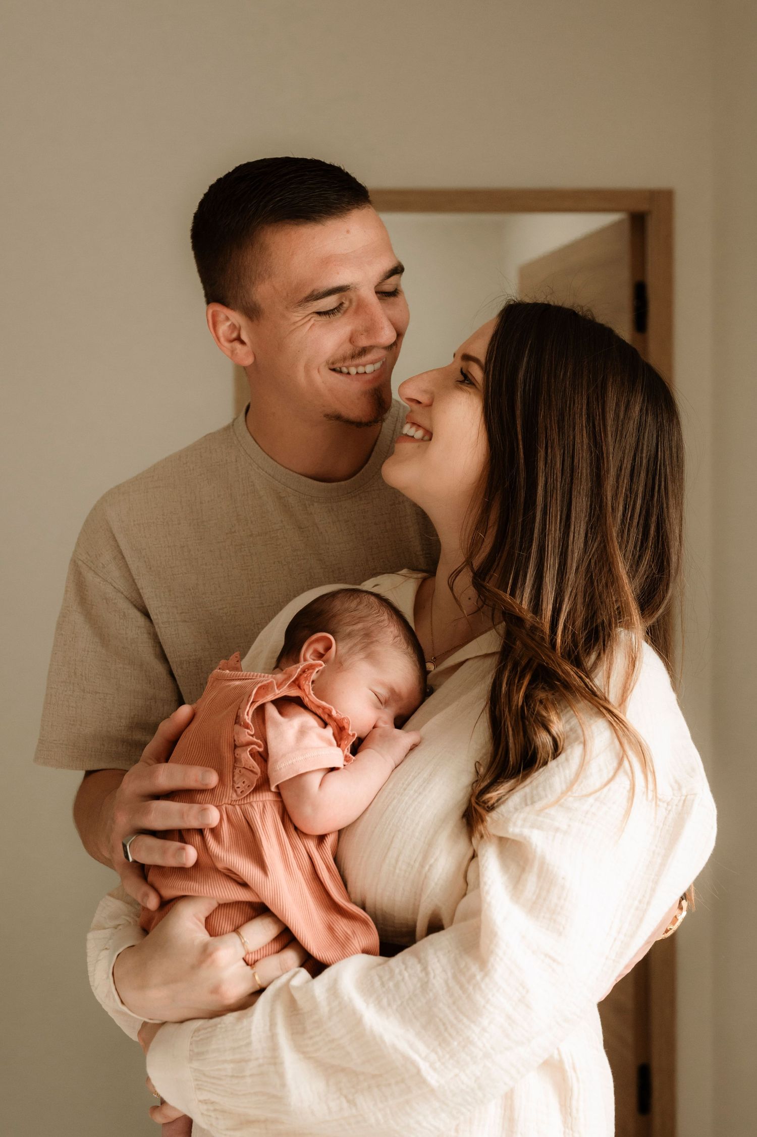 A tender family moment with parents holding their sleeping newborn baby in a neutral-toned indoor setting.