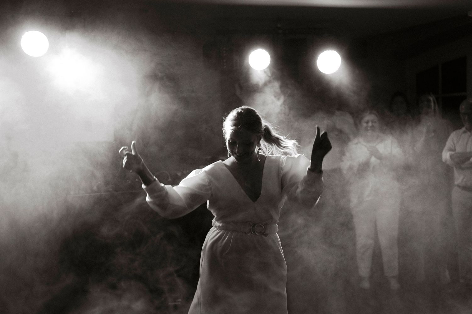 Black and white photo of a woman performing on a smoky stage with spotlights and audience in background.