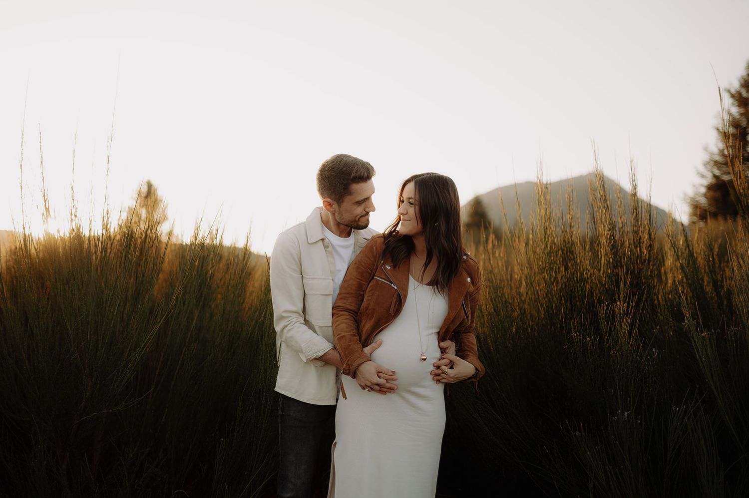 A loving couple embraces in a field of tall grass at golden hour with mountains in the background.