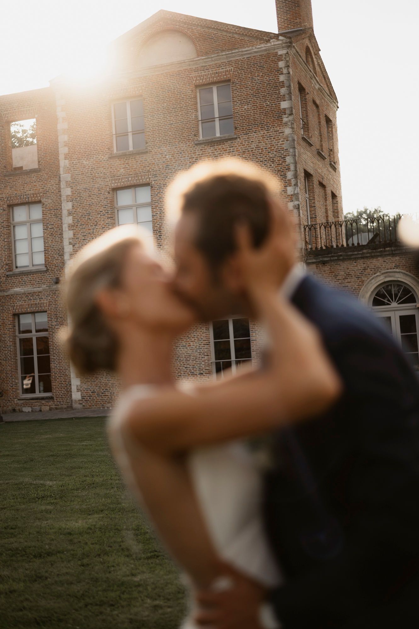 Blurred couple sharing a romantic kiss at golden hour in front of a historic brick mansion at their wedding.