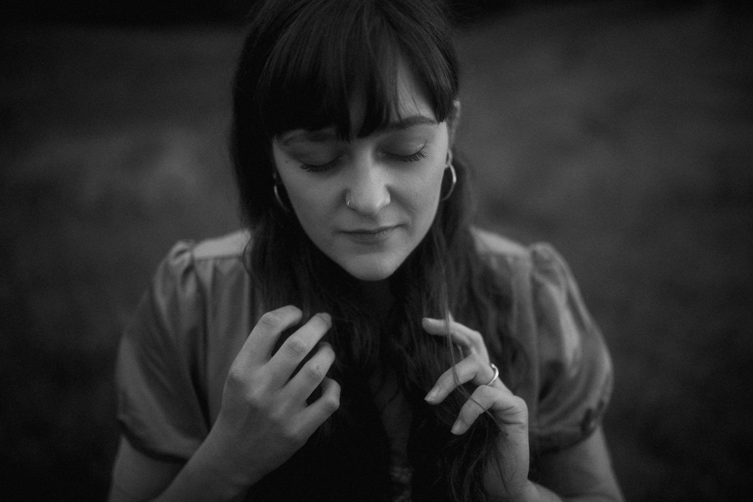 Black and white portrait of someone with dark bangs gazing down while holding their hands near their collar.