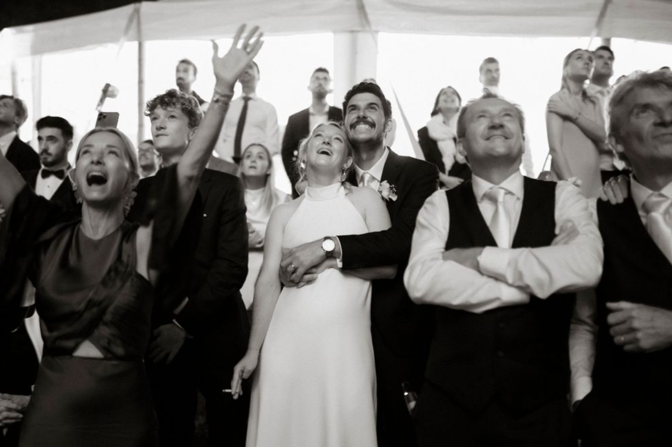 Black and white wedding photo of bride and groom laughing with guests looking up at bouquet toss.