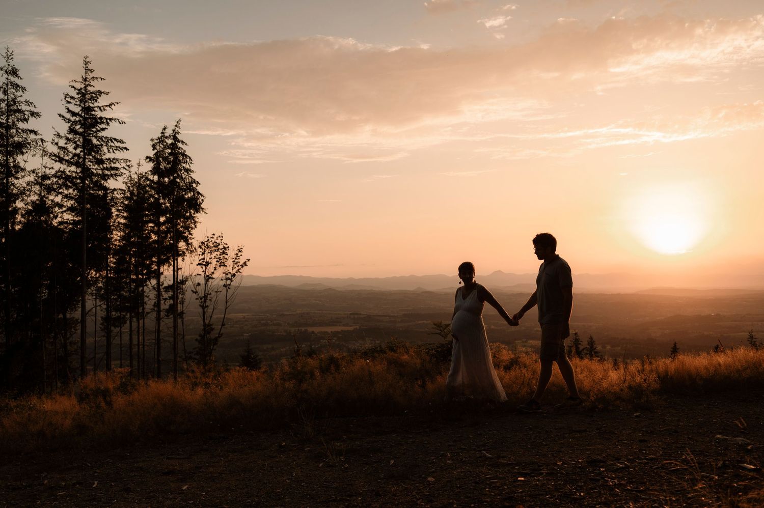 Silhouettes stand together on a hilltop against a warm sunset sky with evergreen trees in the background.