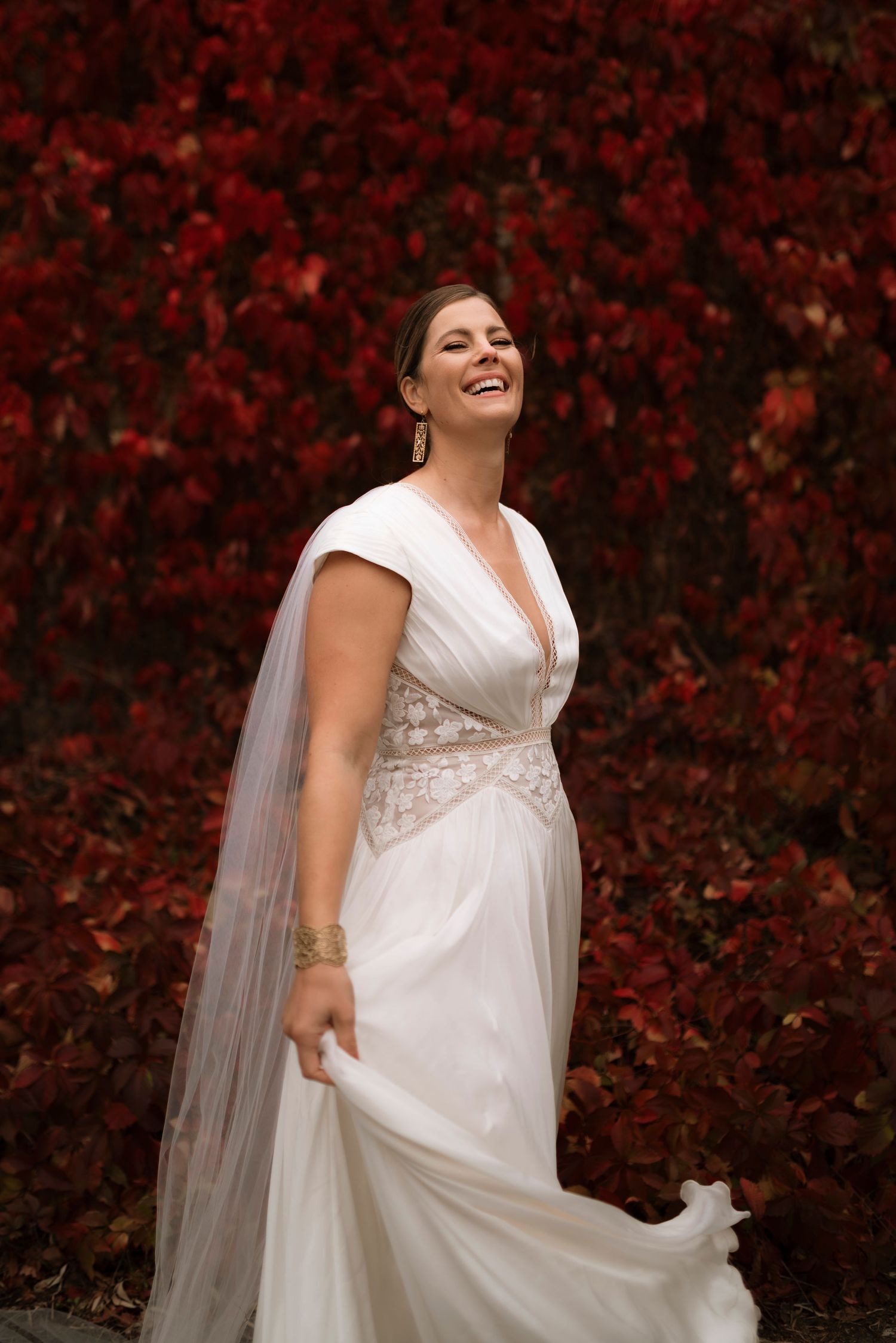 Bride in white gown laughing joyfully against vibrant red autumn foliage backdrop.