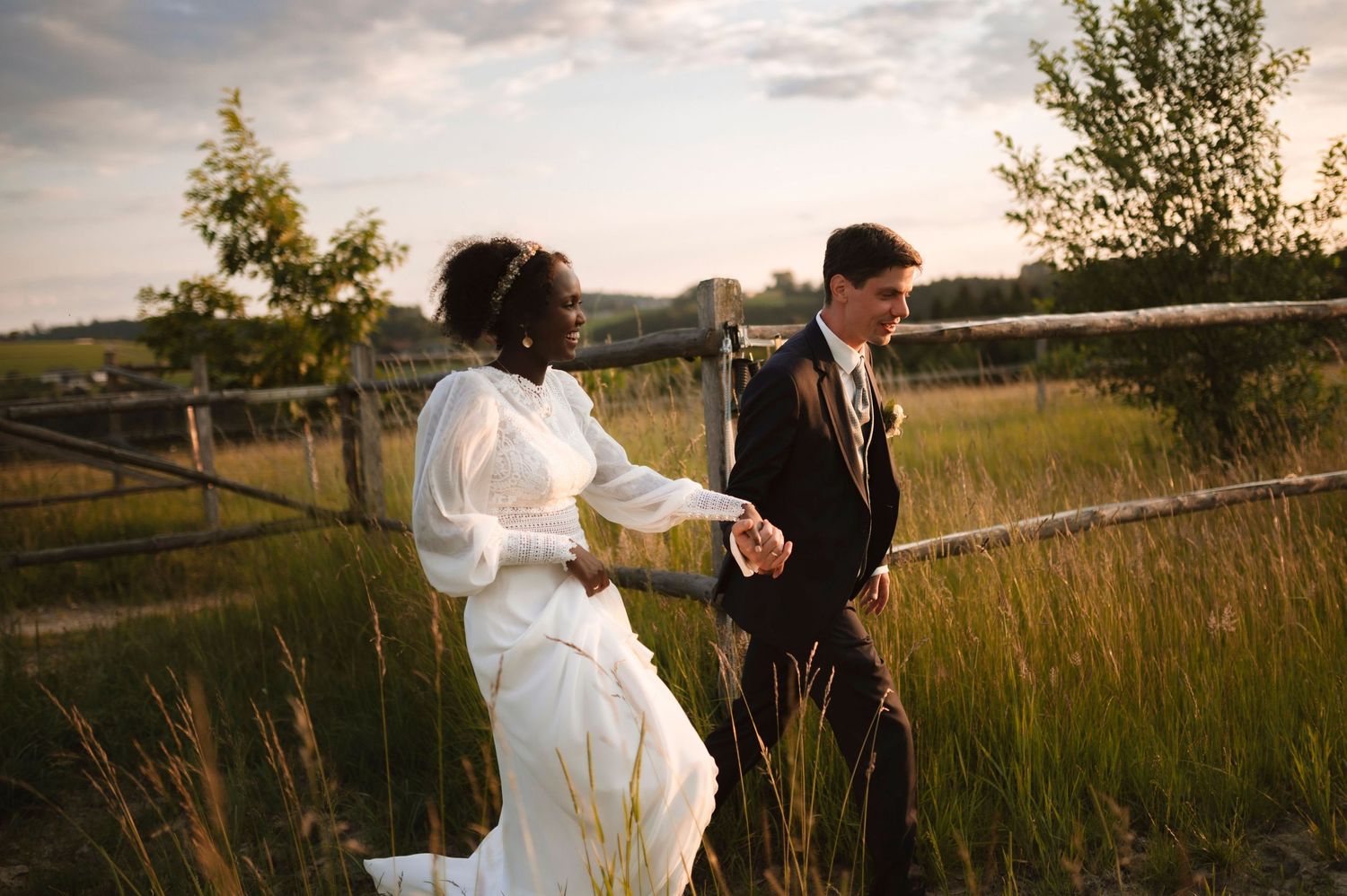 Interracial couple in wedding attire holding hands and walking through a lush green field at golden hour.