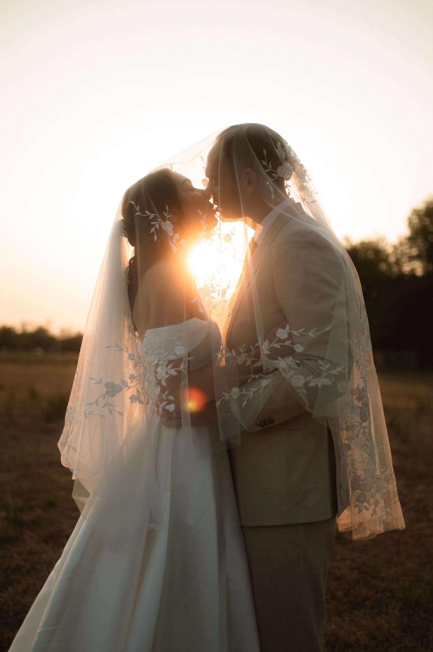 Bride and groom kissing under lace veil at golden sunset in an open field, romantic wedding portrait.