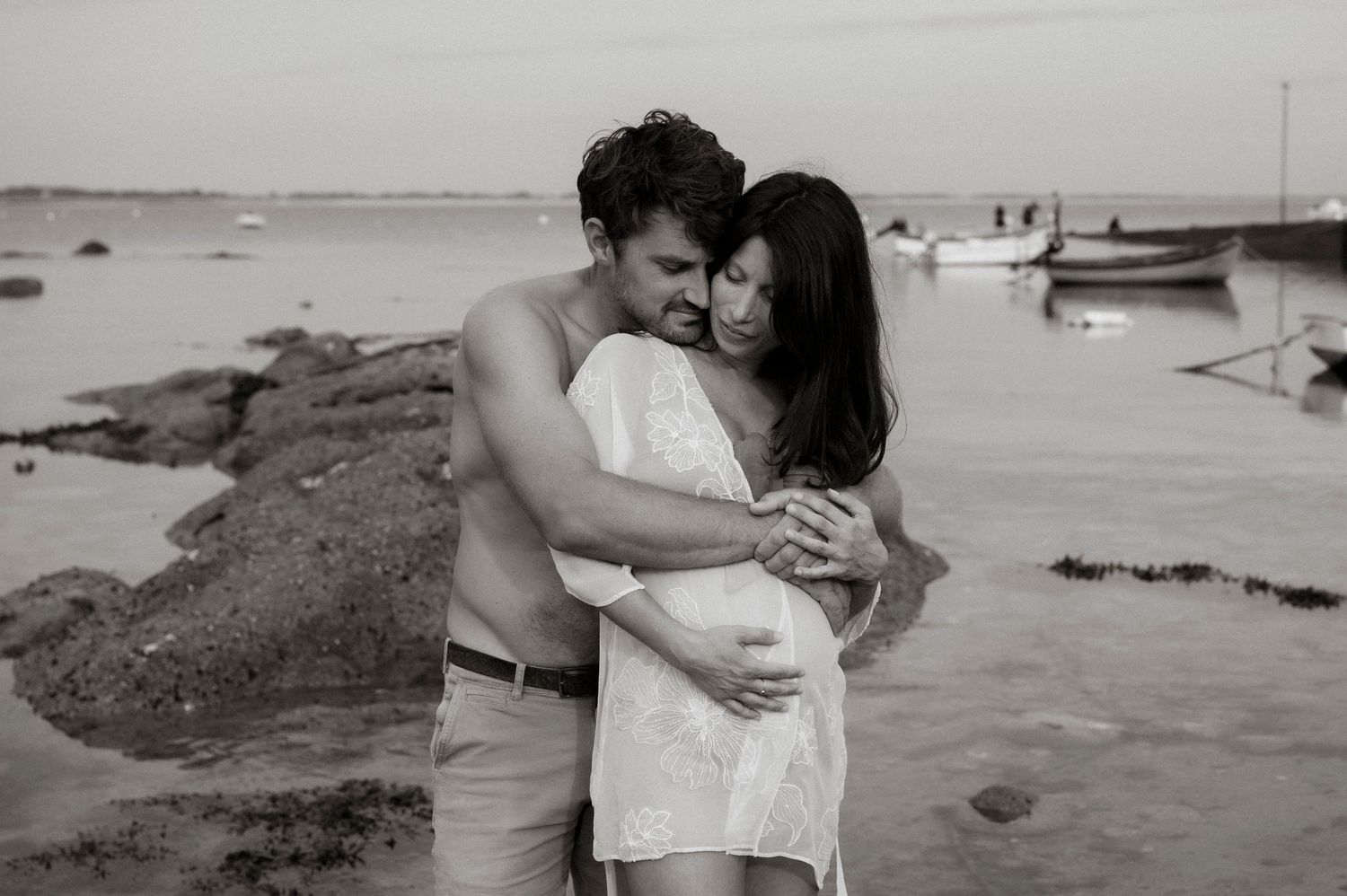 A series of black and white beach engagement photos showing a couple embracing on a rocky coastline at sunset.