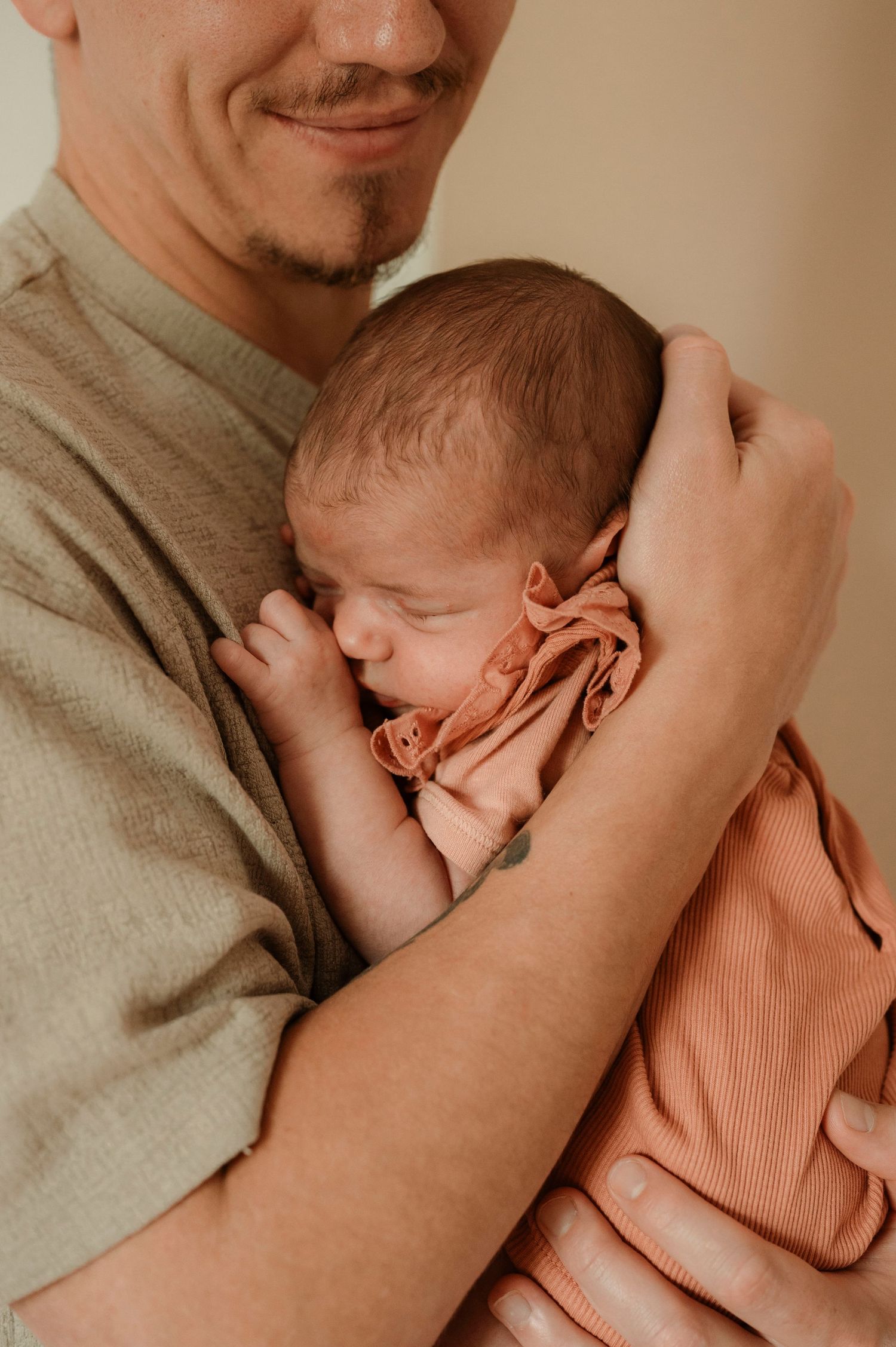 A sleeping newborn baby in a peachy pink outfit is cradled lovingly against someone wearing a grey shirt.