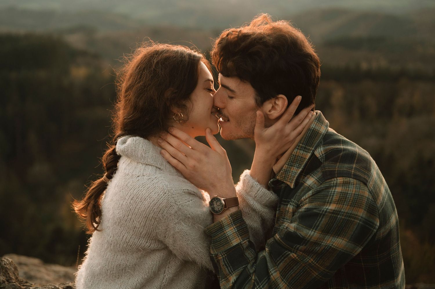 A couple shares a tender kiss at sunset in nature, with golden light illuminating them against a mountain backdrop.