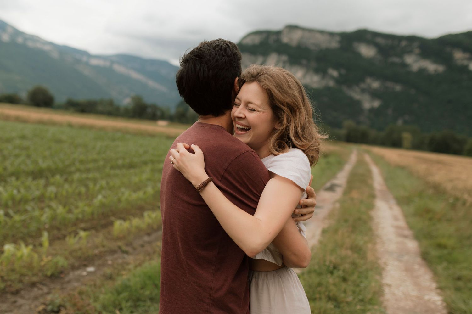 A couple embraces joyfully on a dirt path with mountains and green fields in the scenic countryside background.