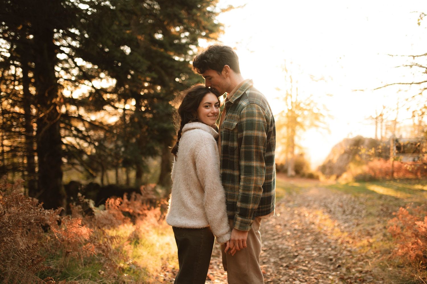 A couple embraces on a sunlit autumn path surrounded by golden ferns and tall evergreen trees at sunset.