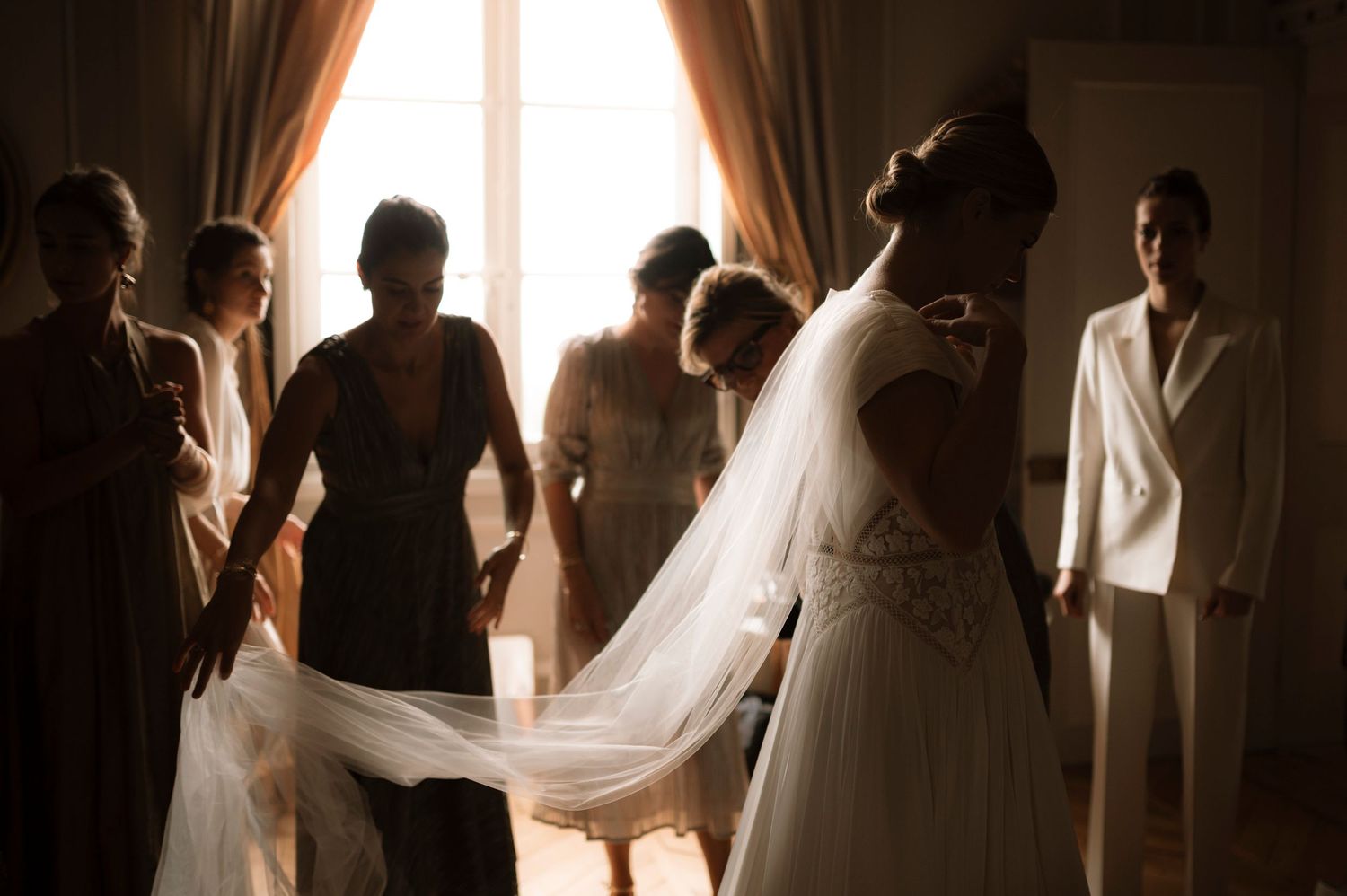 Bride in white gown and veil surrounded by bridesmaids and guests in elegant room during wedding preparation.