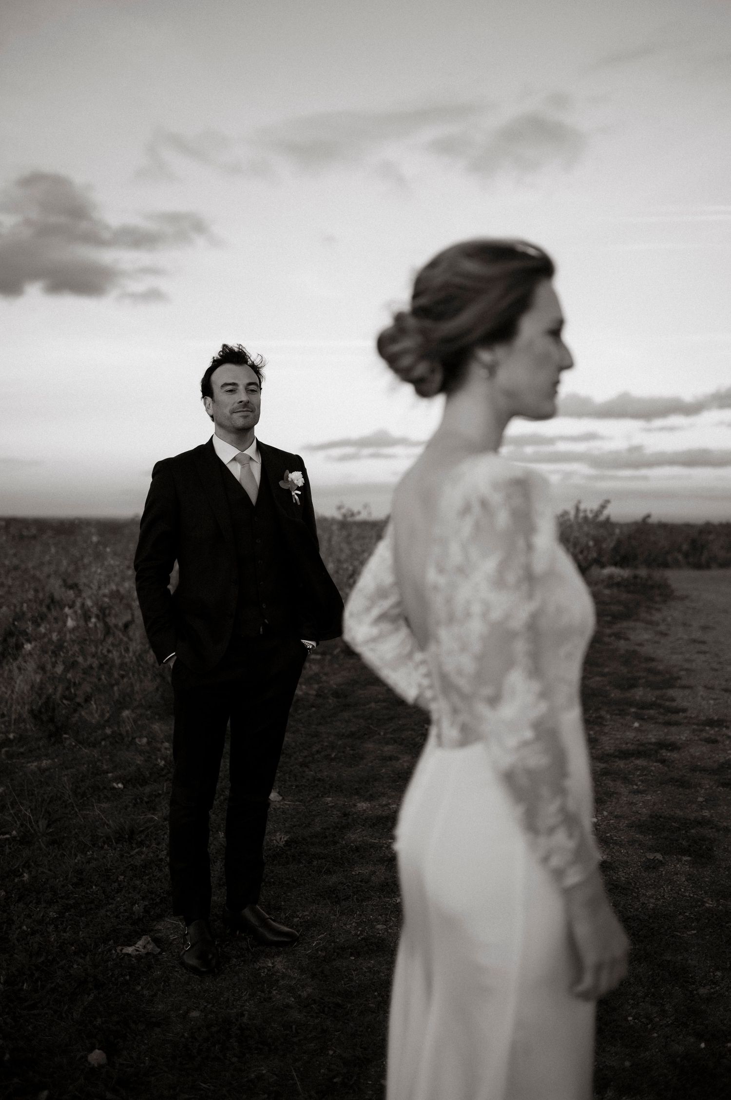 Black and white wedding photo of groom gazing at bride in lace dress against dramatic sunset sky.