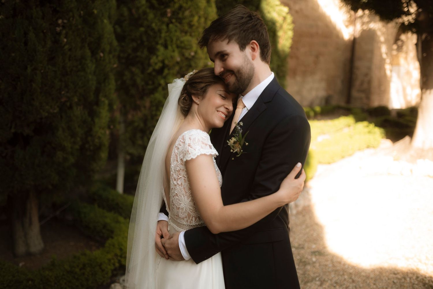 Newlywed couple embraces tenderly on a sunlit garden path during their wedding celebration.