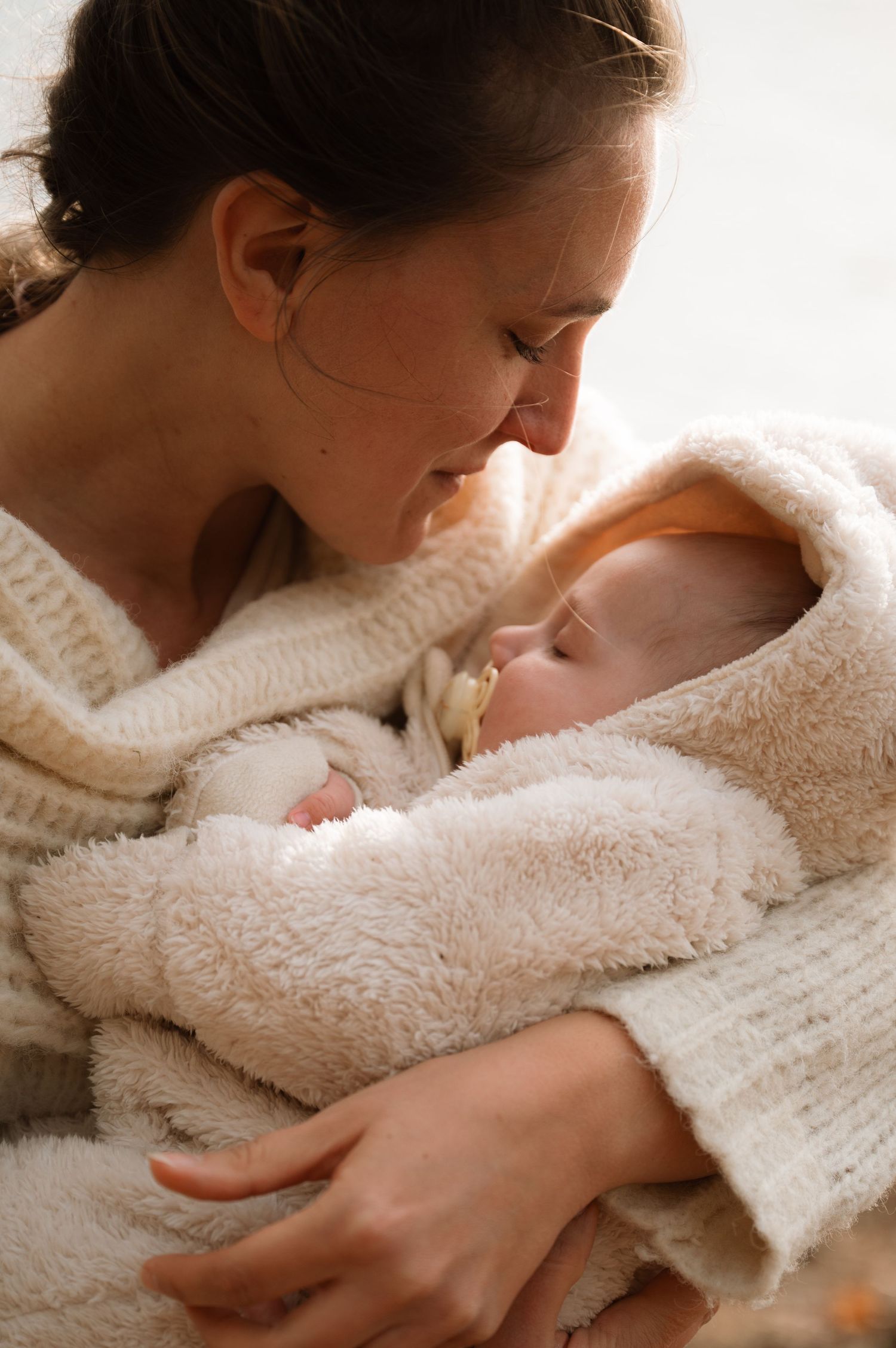 A tender moment between mother and baby wrapped in soft white blankets in warm natural lighting.