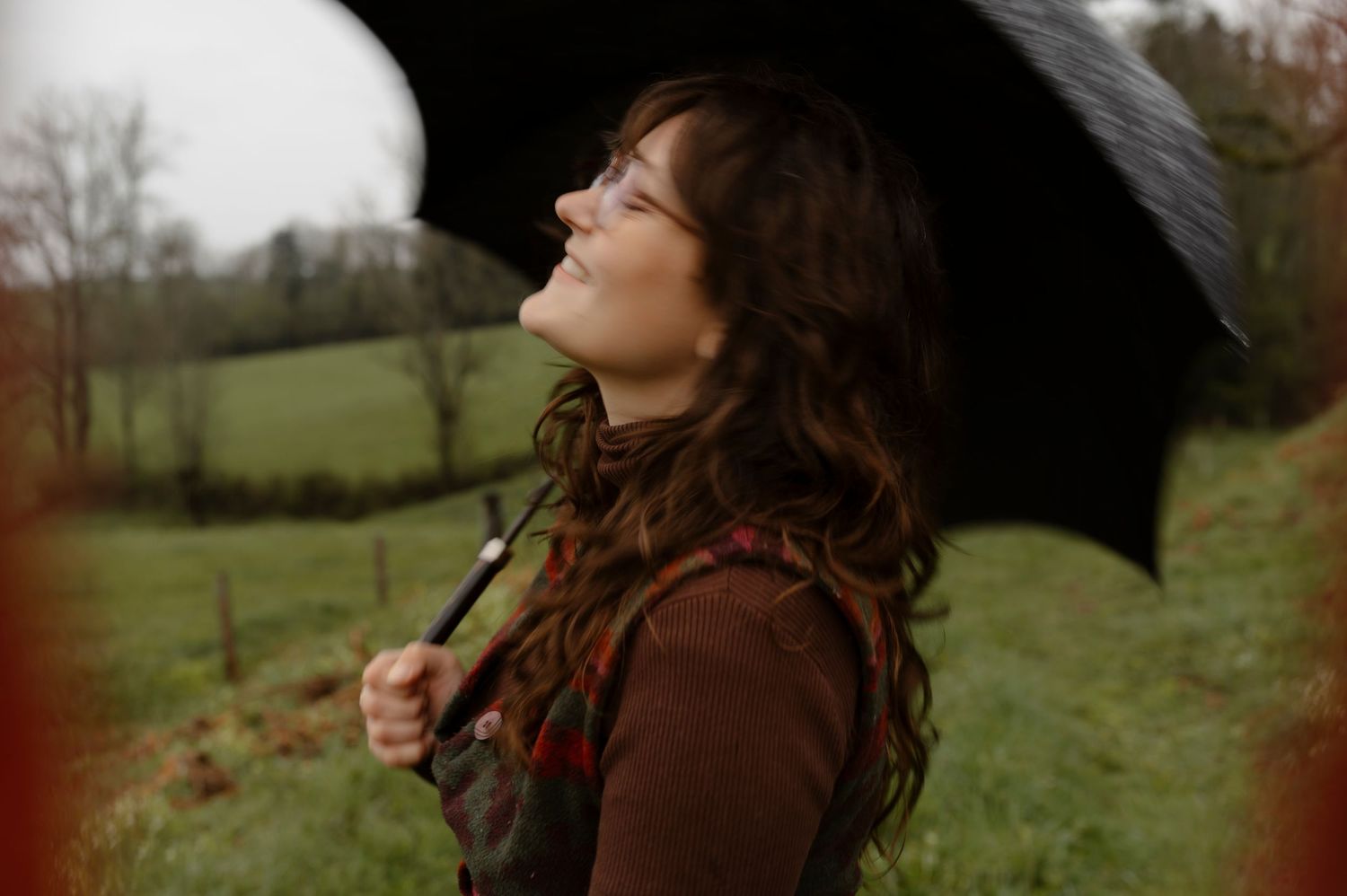 Person in brown sweater holding black umbrella stands in profile against a green countryside on an overcast day.