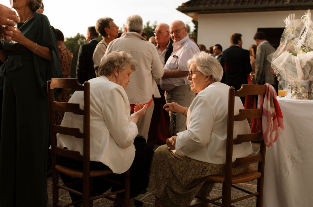 Two elderly women in white jackets sit on wooden chairs conversing at an outdoor social gathering or wedding reception.