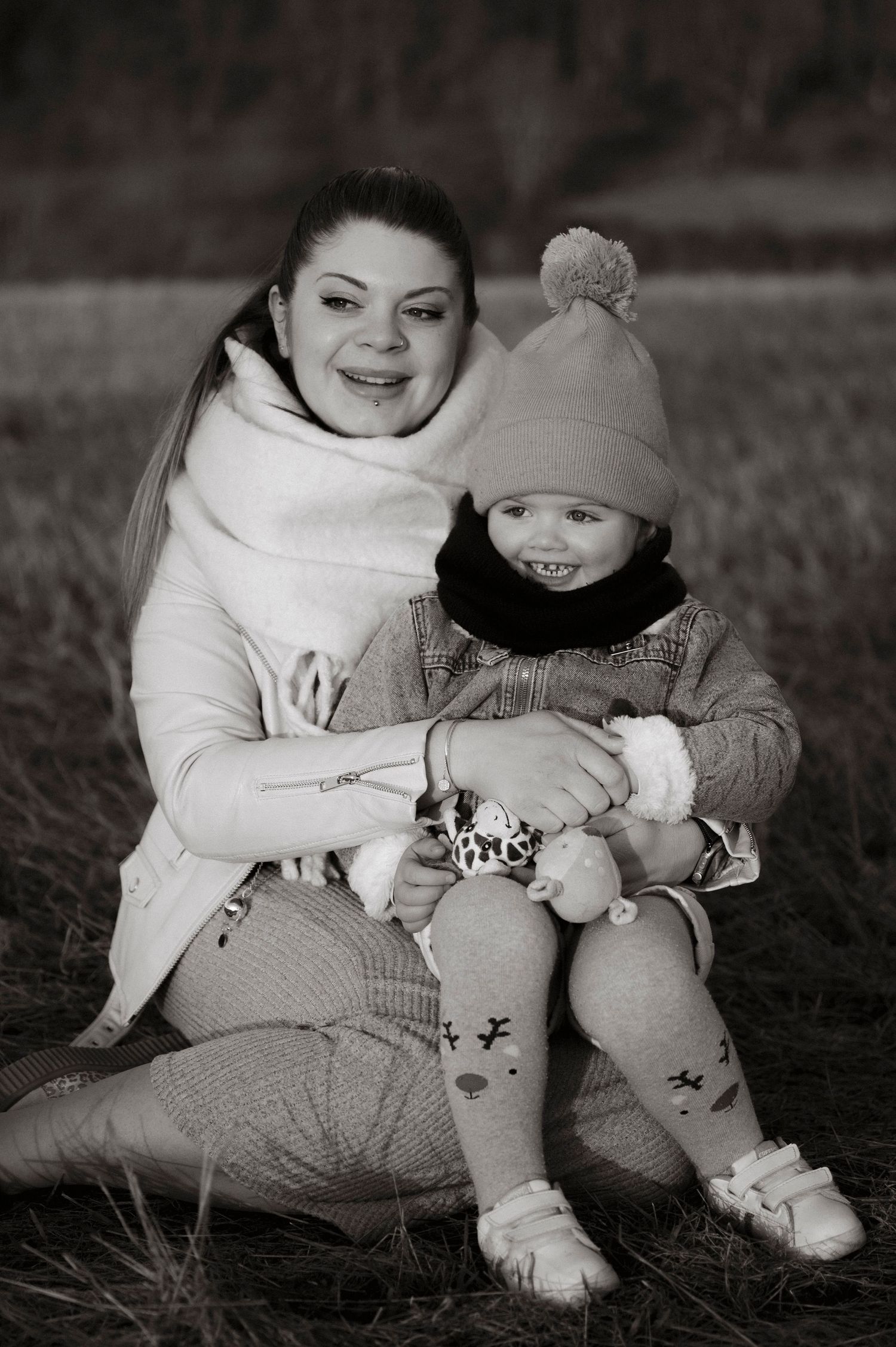 Black and white portrait of parent and child sitting outdoors in winter clothes together on grass.