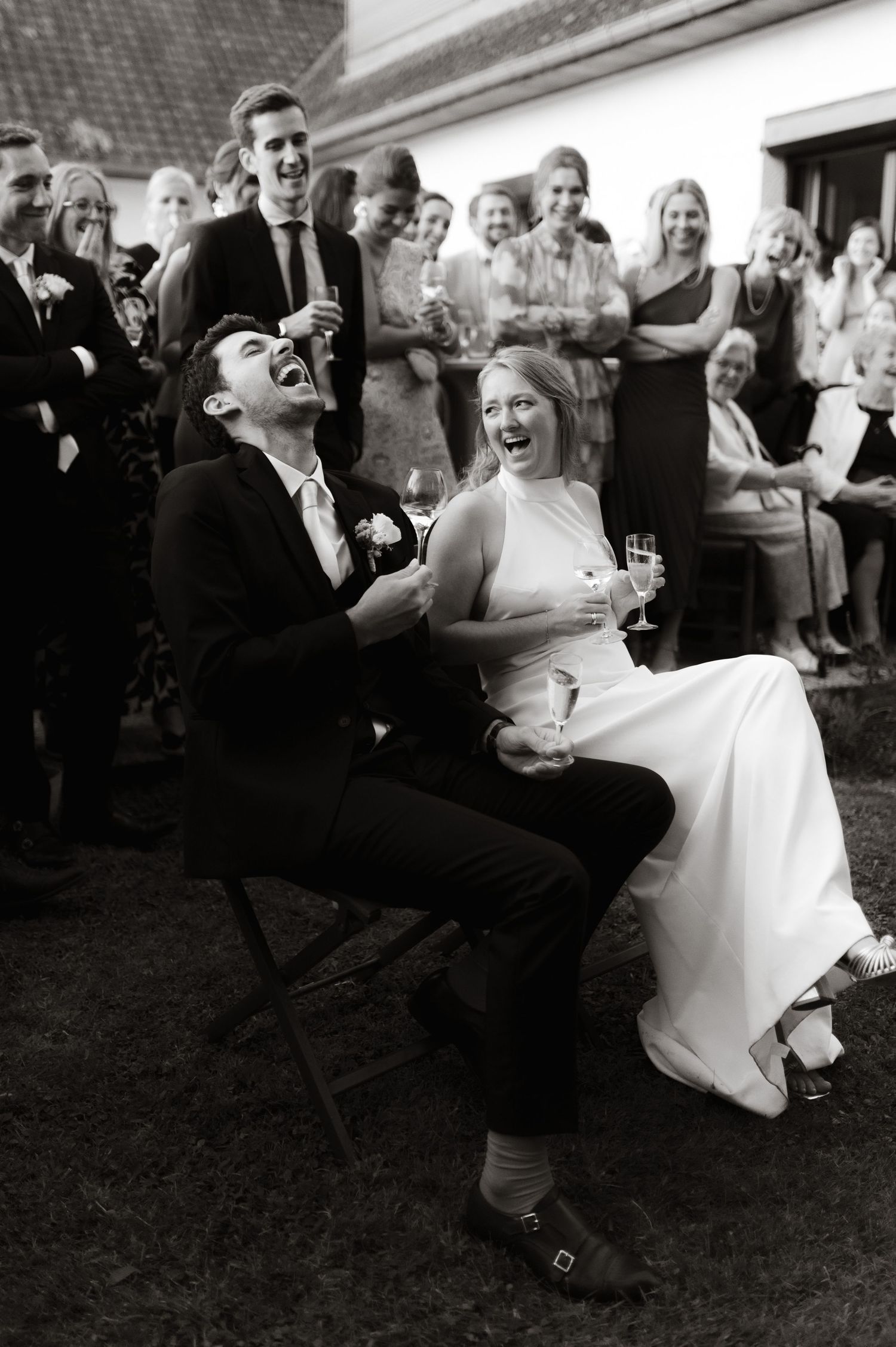 Black and white photo of laughing bride and groom seated at outdoor wedding reception with guests watching.
