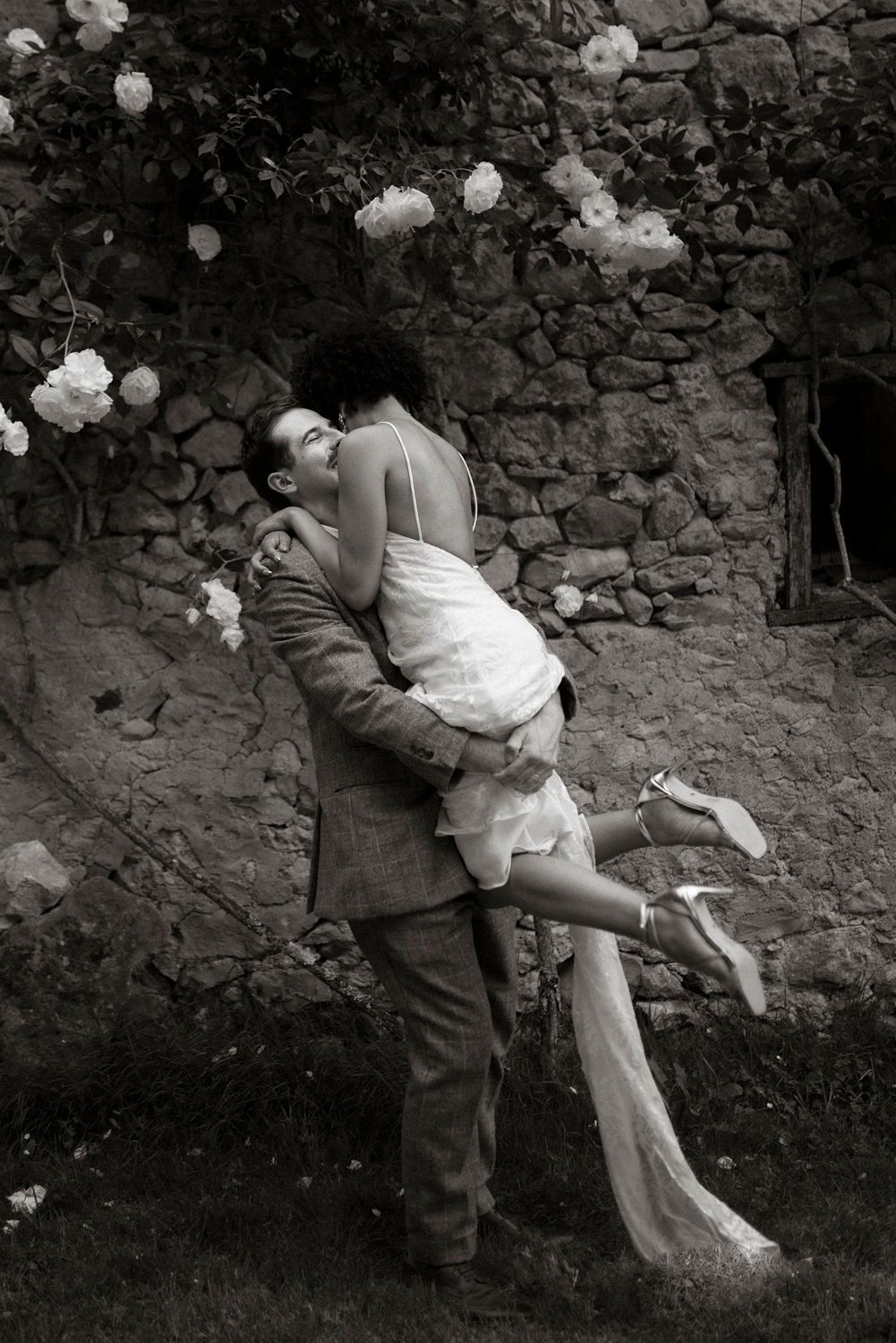 Black and white photo of a groom lifting his bride near a stone wall with blooming roses.