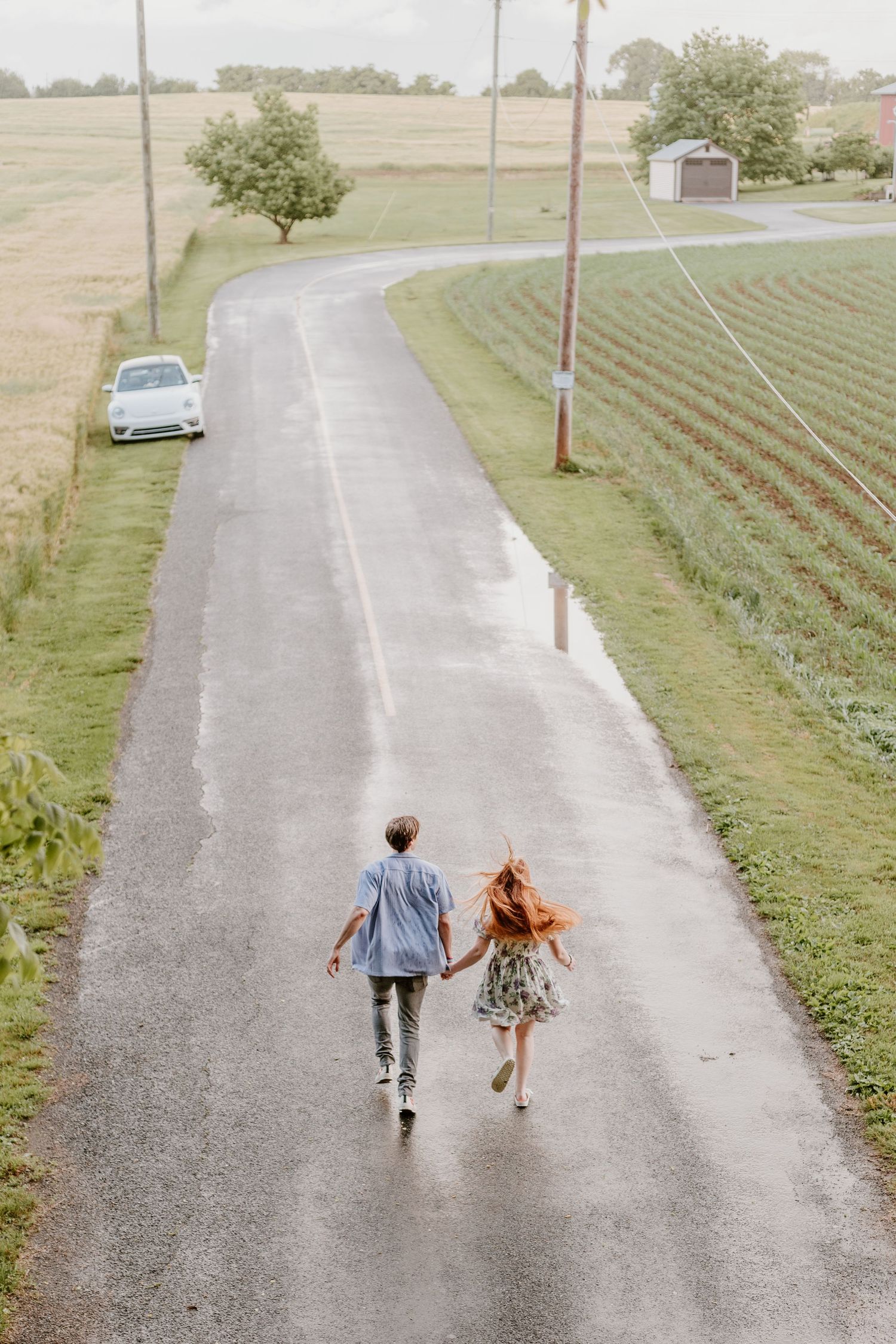 A couple runs playfully down a rural road with fields and a car in the background.