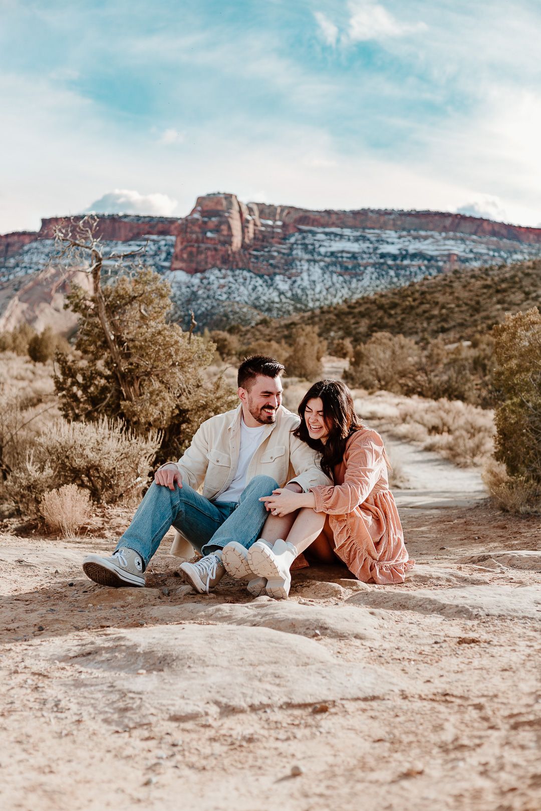 A couple sits on desert ground with scenic red rock formations in the background.