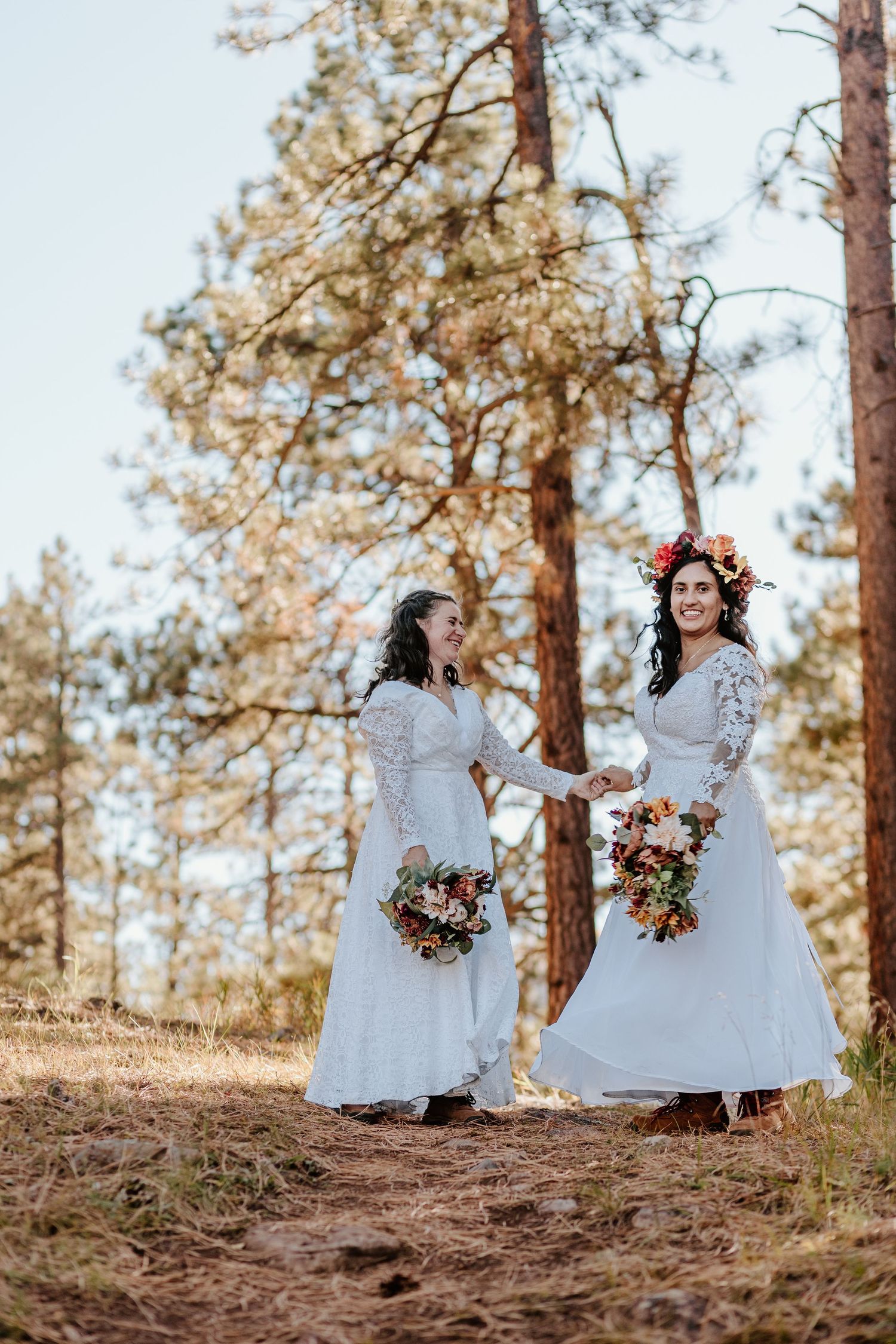 Two people in white lace dresses hold floral bouquets while standing among tall pine trees.
