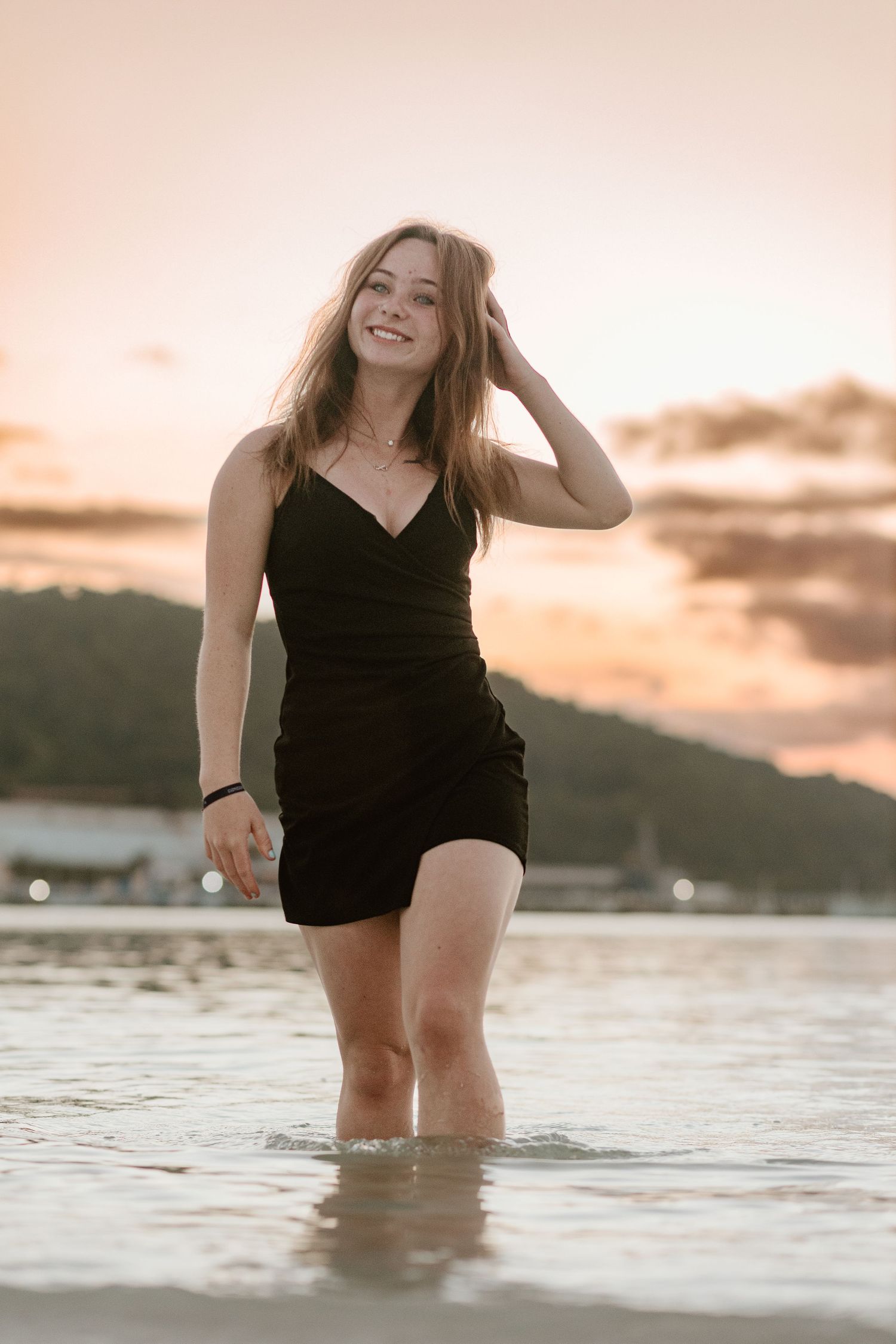 A person wearing a black romper stands in shallow water during sunset.