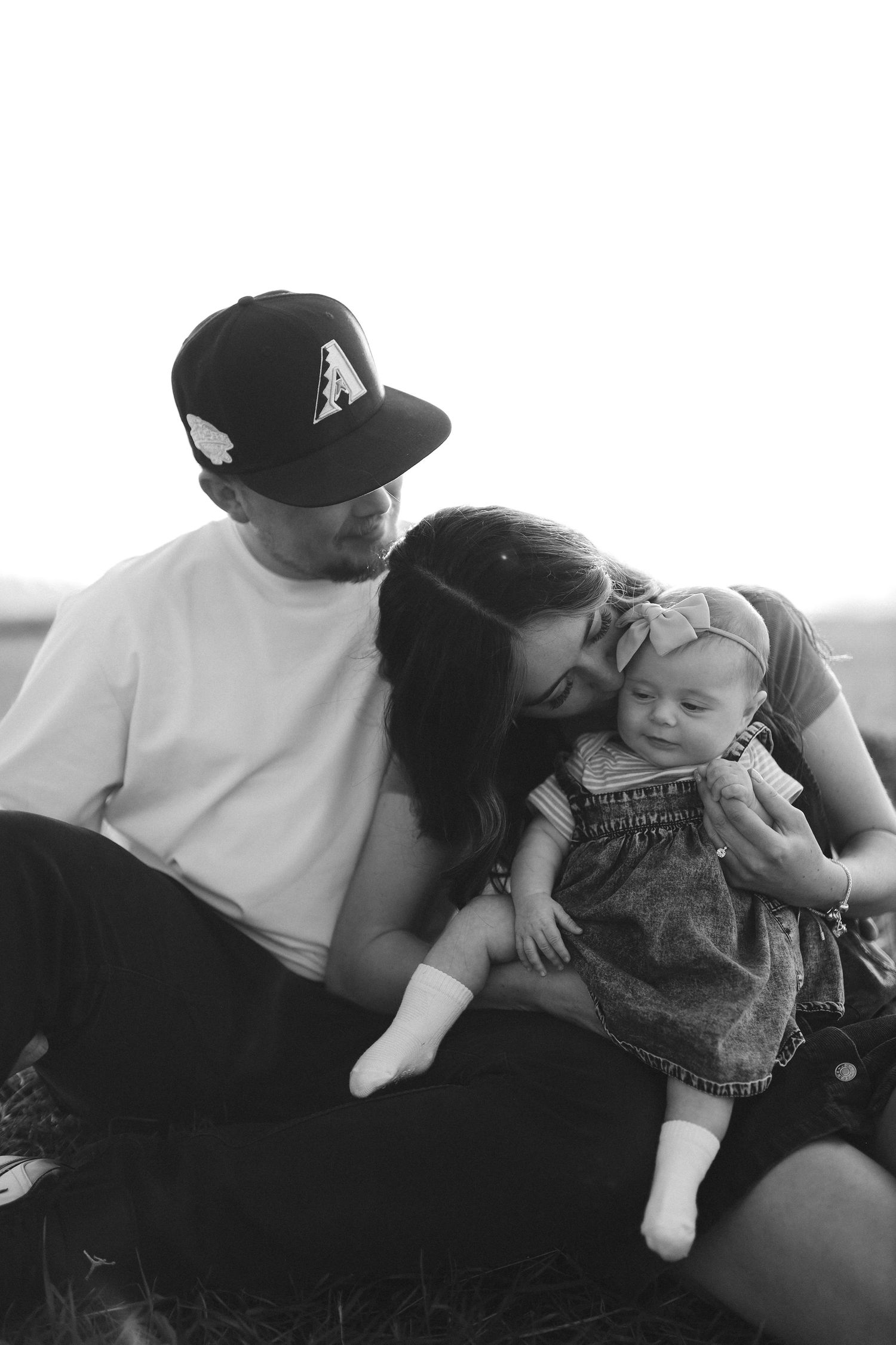 A black and white photo of parents sitting together, lovingly kissing their newborn baby outdoors.