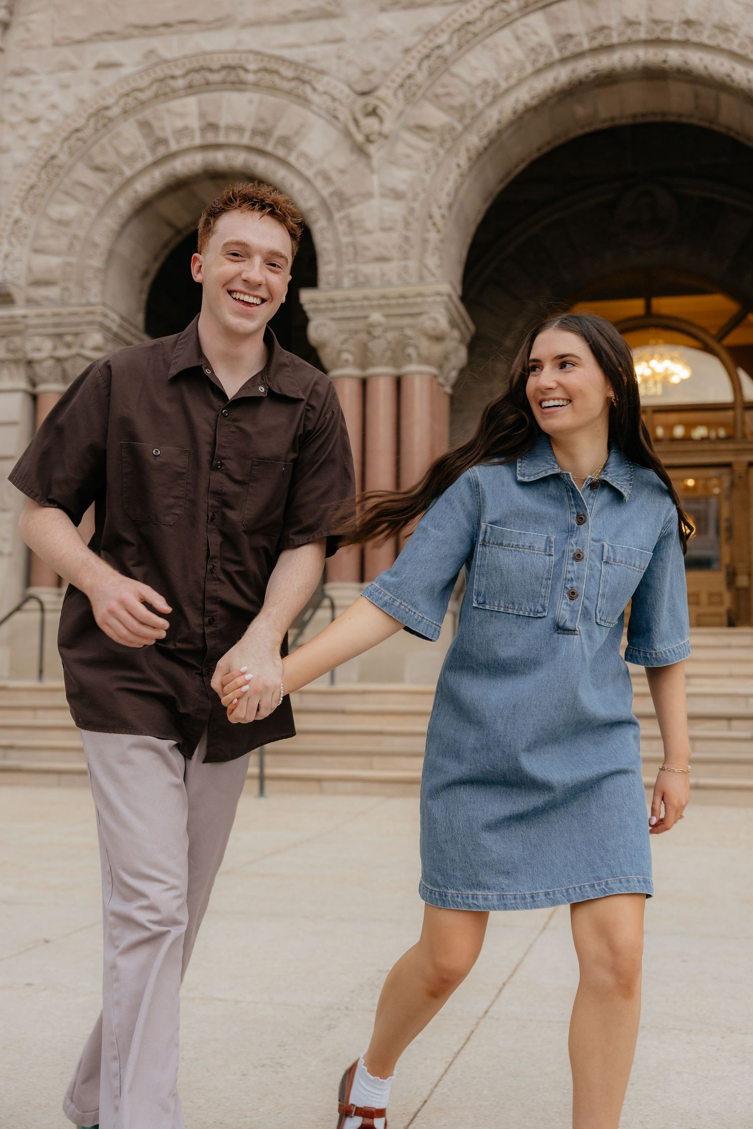 A joyful couple walks hand-in-hand, laughing together outside a grand stone archway building.