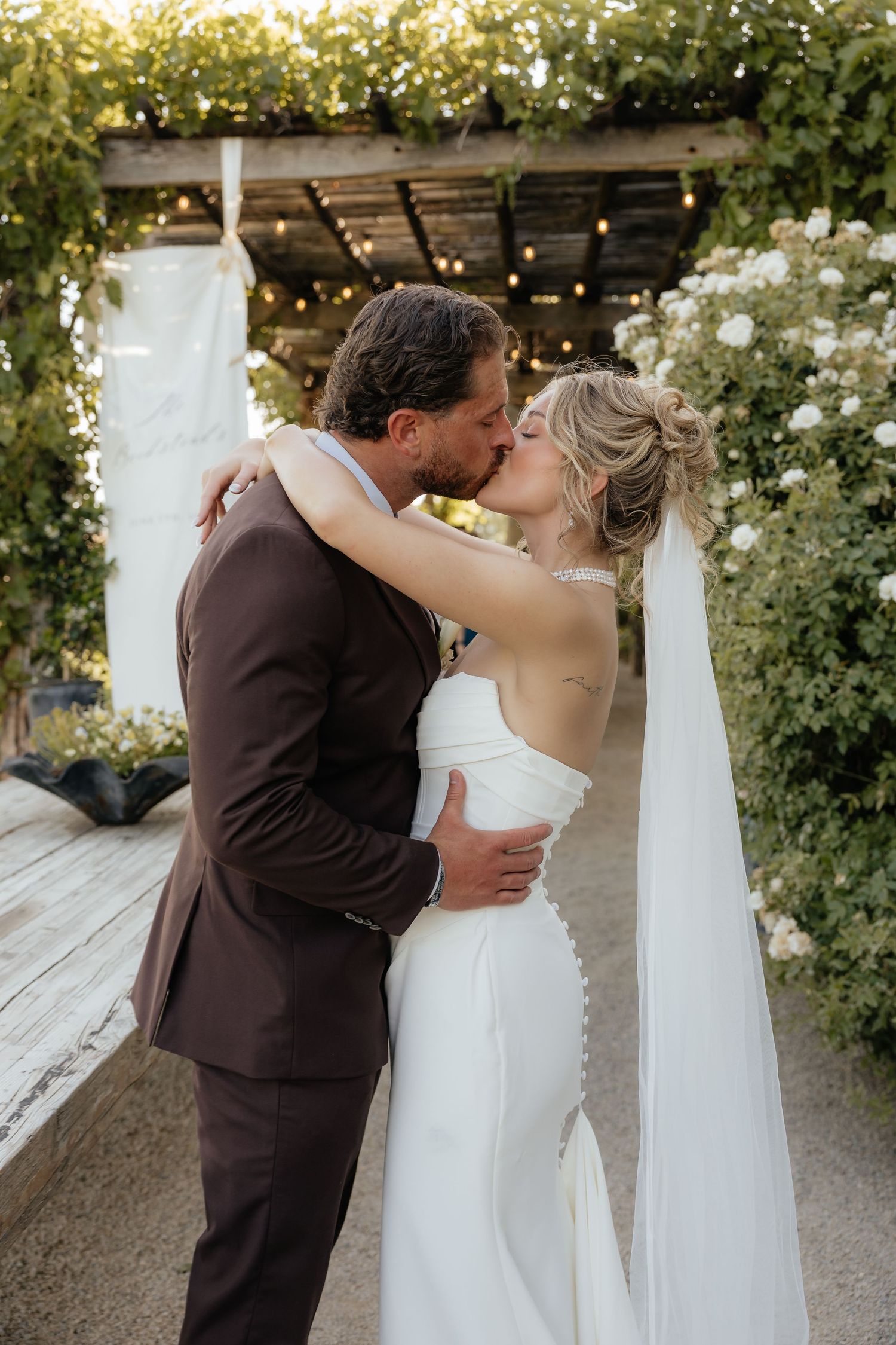 Bride and groom kissing under a vine-covered pergola with string lights at outdoor wedding.