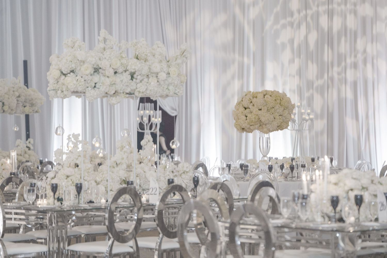 White floral arrangements and crystal chandeliers adorn an elegant wedding reception venue with draped white fabric decor.