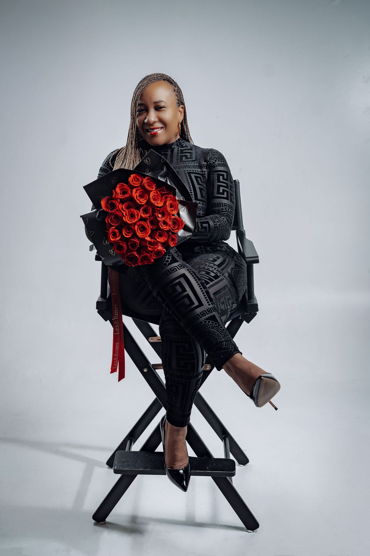 A person in a black patterned outfit sits on a director's chair holding a bouquet of red roses against a gray background.