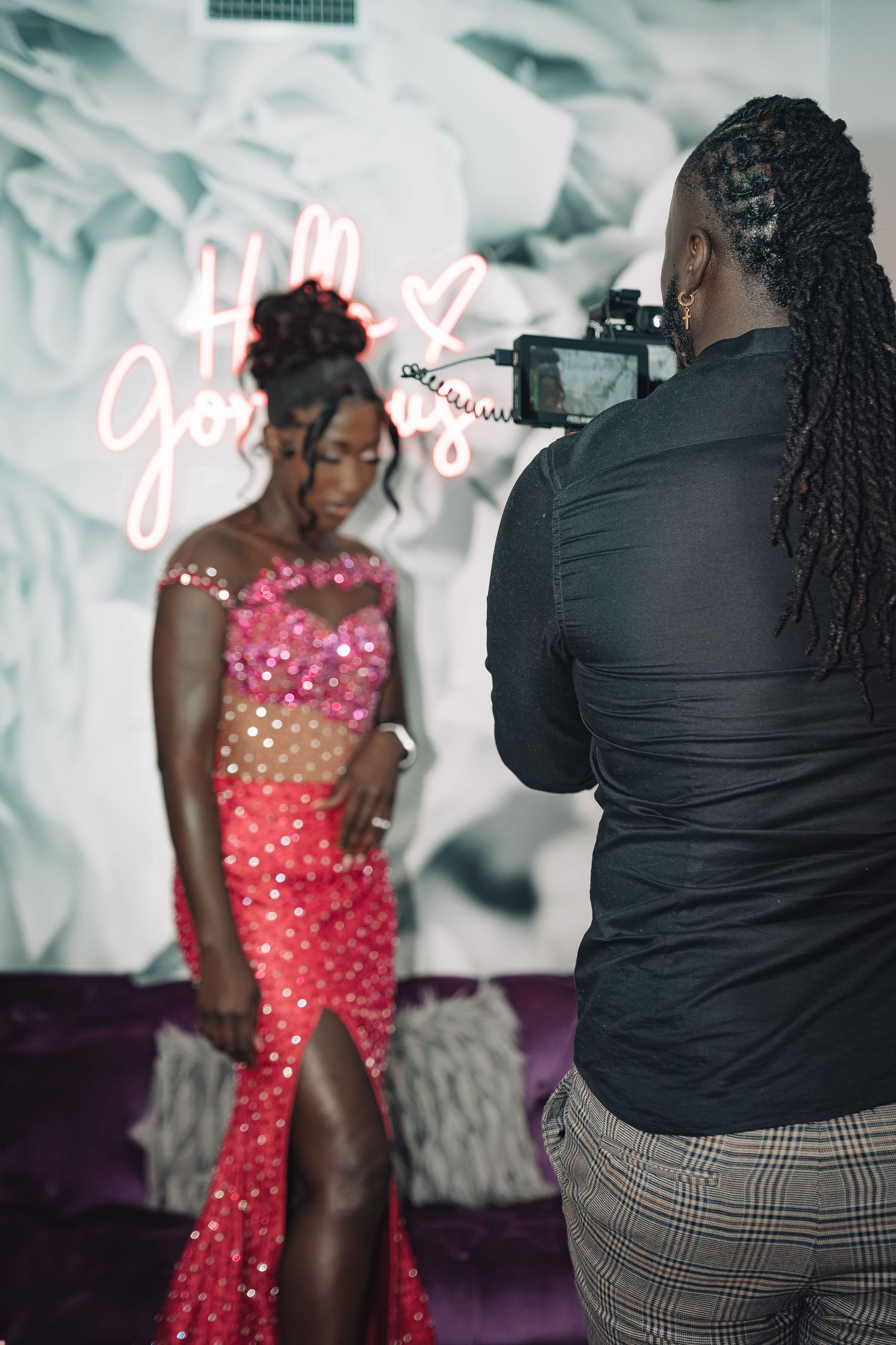 Photographer captures model in sparkly red evening gown with thigh slit against white backdrop with neon sign.