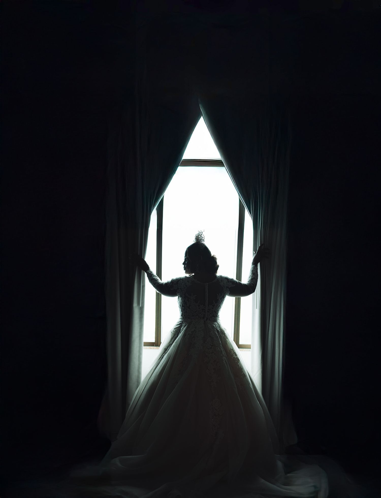 A silhouetted figure in a ballgown dress stands against a bright window between flowing curtains in a dark room.