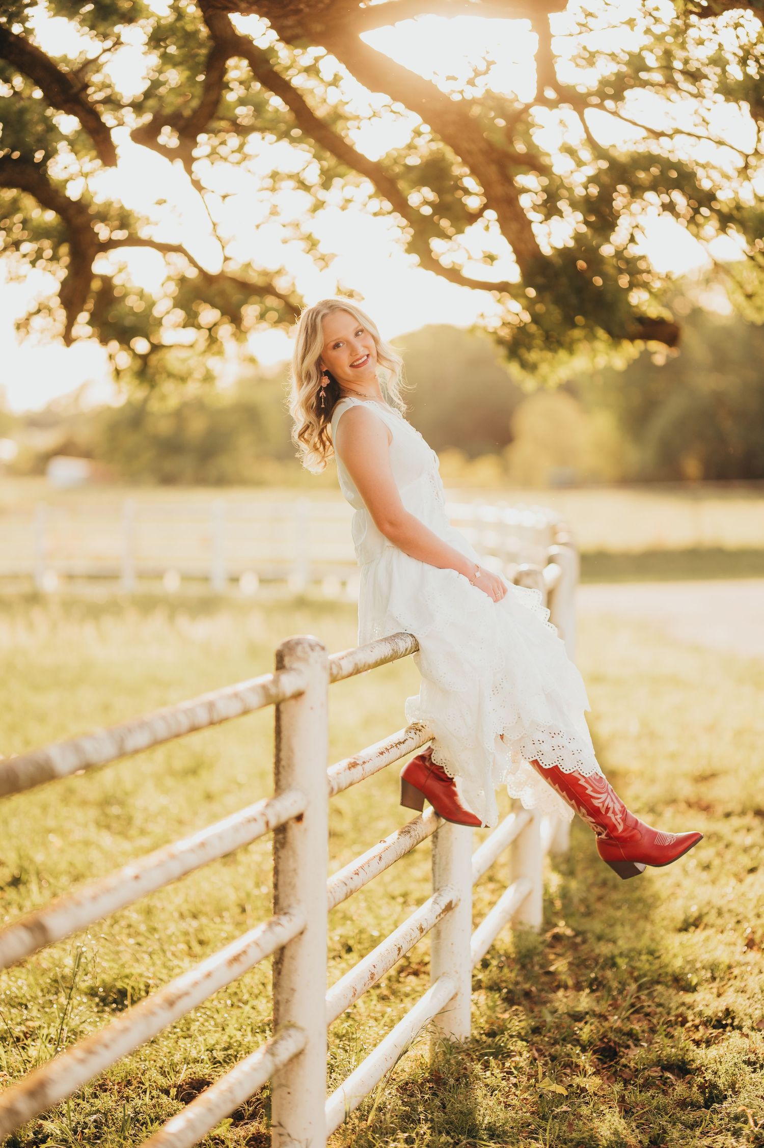 East Texas senior girl in a white dress and red cowboy boots posing by a fence during golden hour