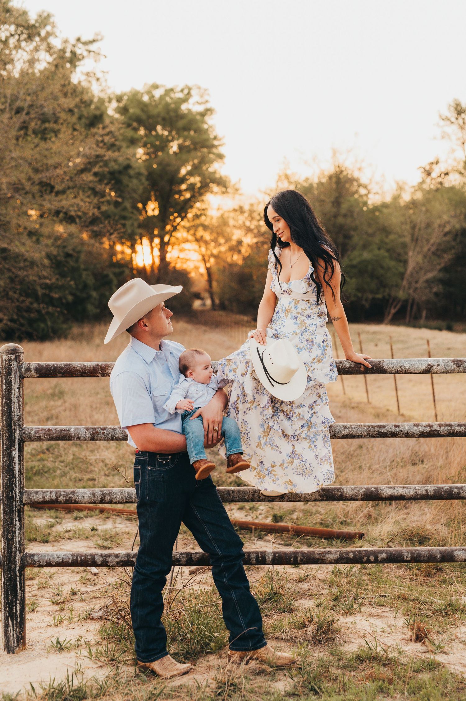 Family portrait of a mother sitting in a fence while the father holds their baby during an outdoor family photography session in East Texas
