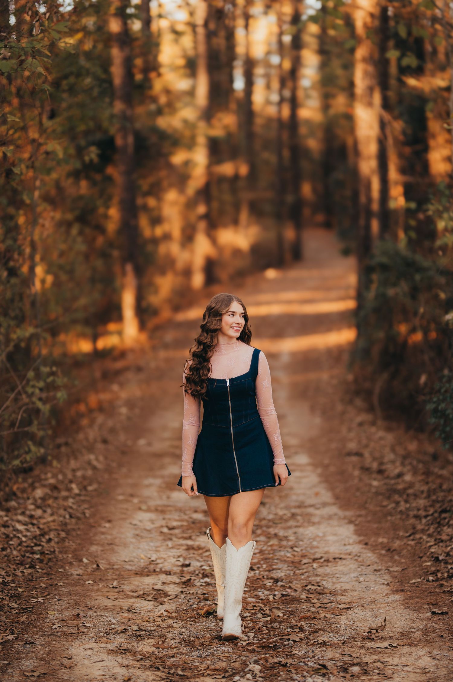 East Texas senior girl in a navy dress and white cowboy boots walking through a wooded path at golden hour