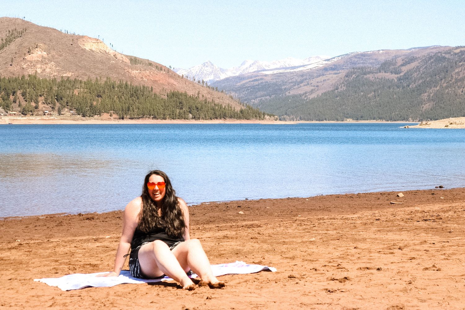 Person relaxing on blanket by scenic mountain lake on sunny day.