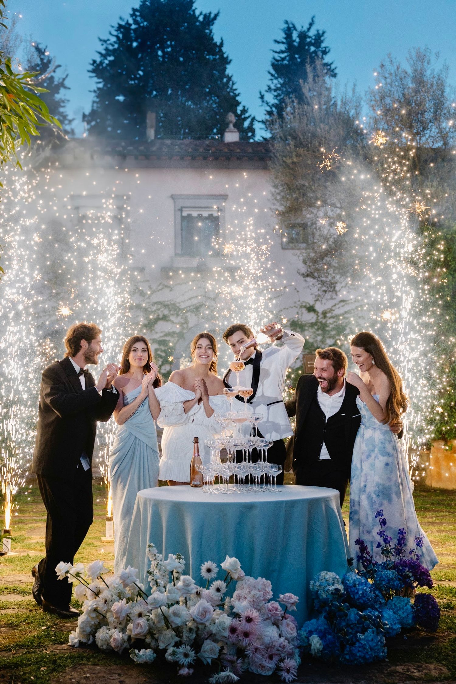 Wedding party celebrating with champagne tower surrounded by dramatic sparkler fountains at dusk.