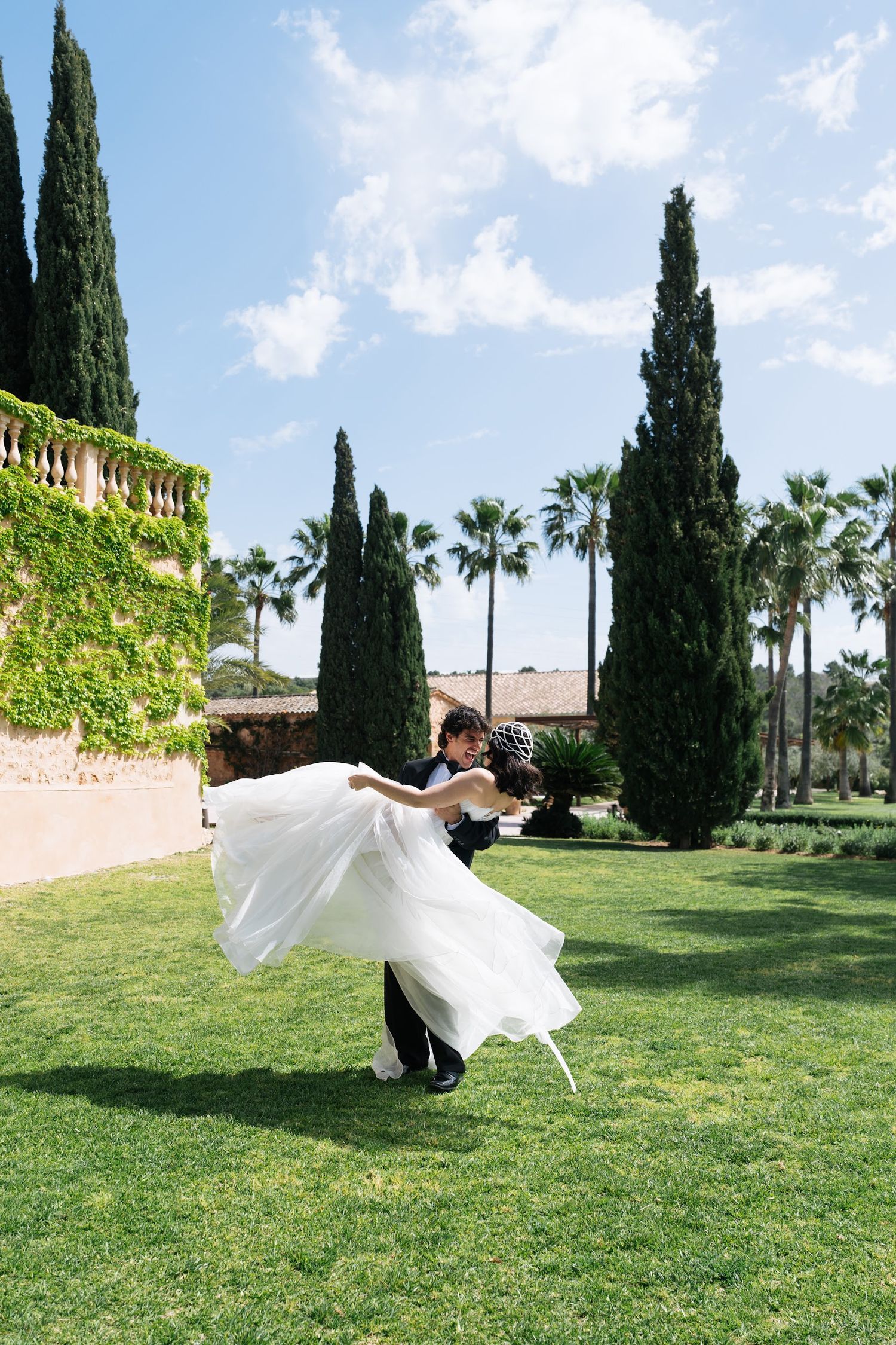 Groom dips bride in flowing white gown on sunny green lawn surrounded by cypress and palm trees.